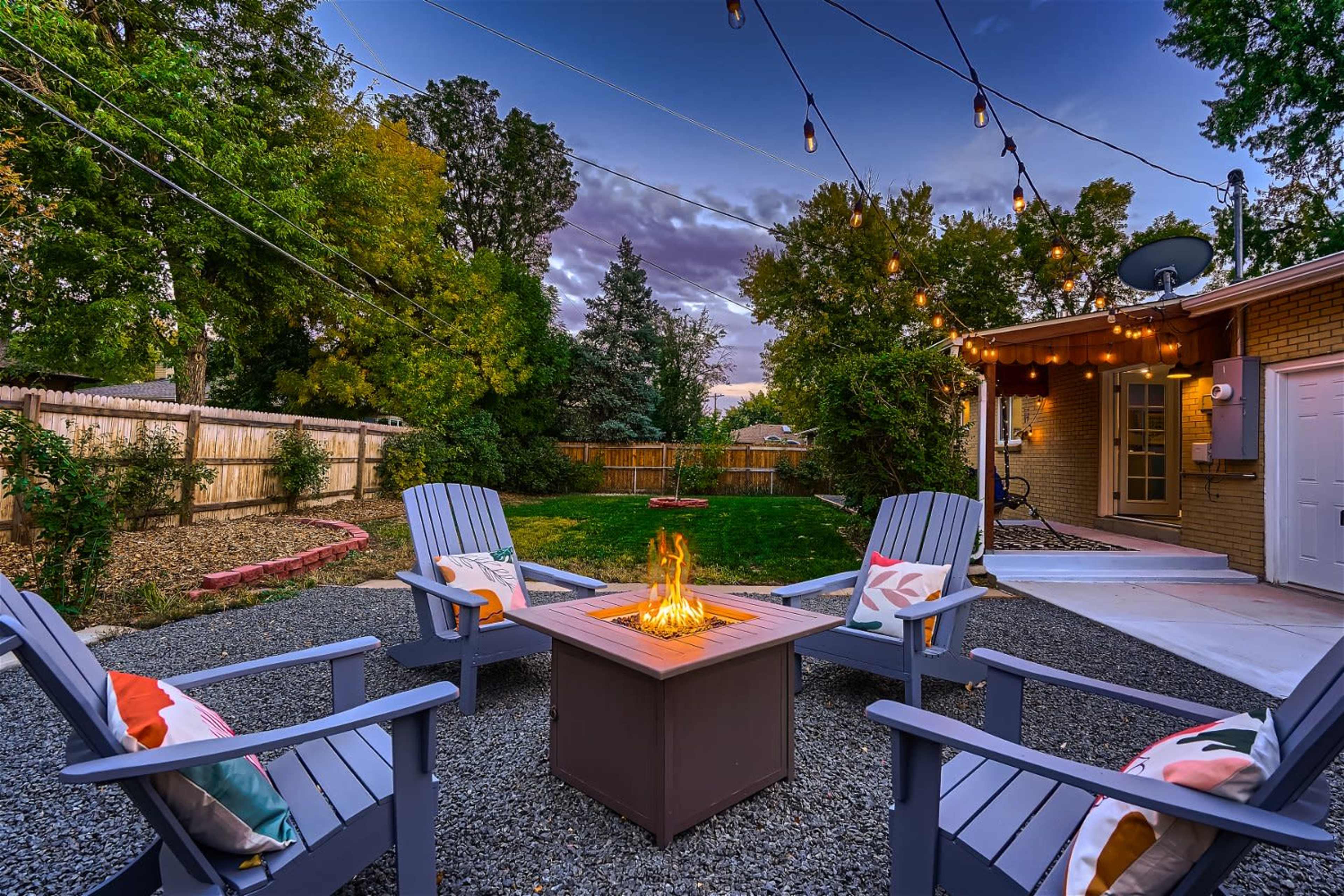 A cozy outdoor seating area features four chairs arranged around a fire pit, with string lights overhead and a grass lawn in the background.