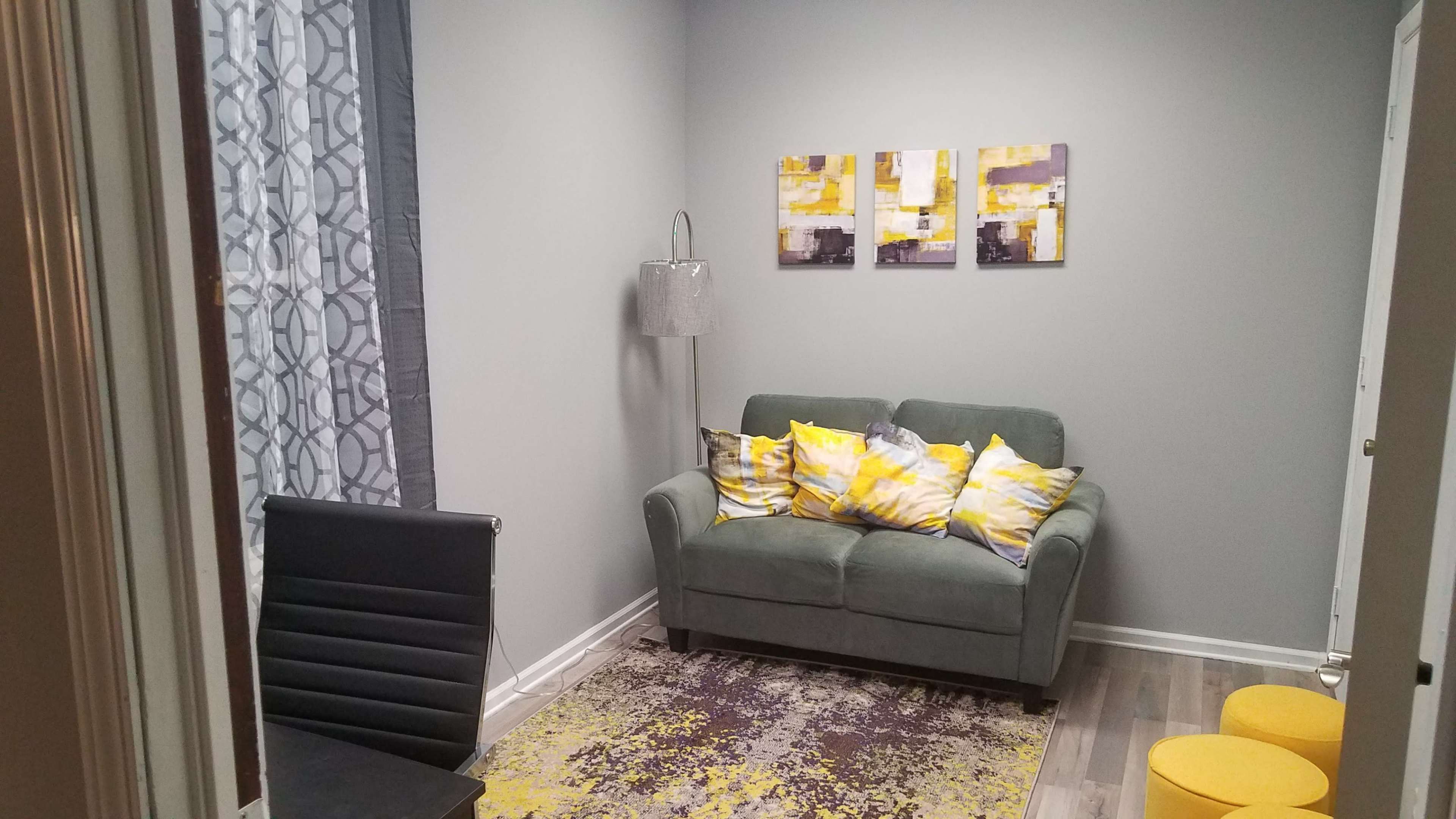 A small room with a green sofa adorned with yellow and gray cushions, a patterned rug on the floor, yellow stools, and abstract artwork on the wall.