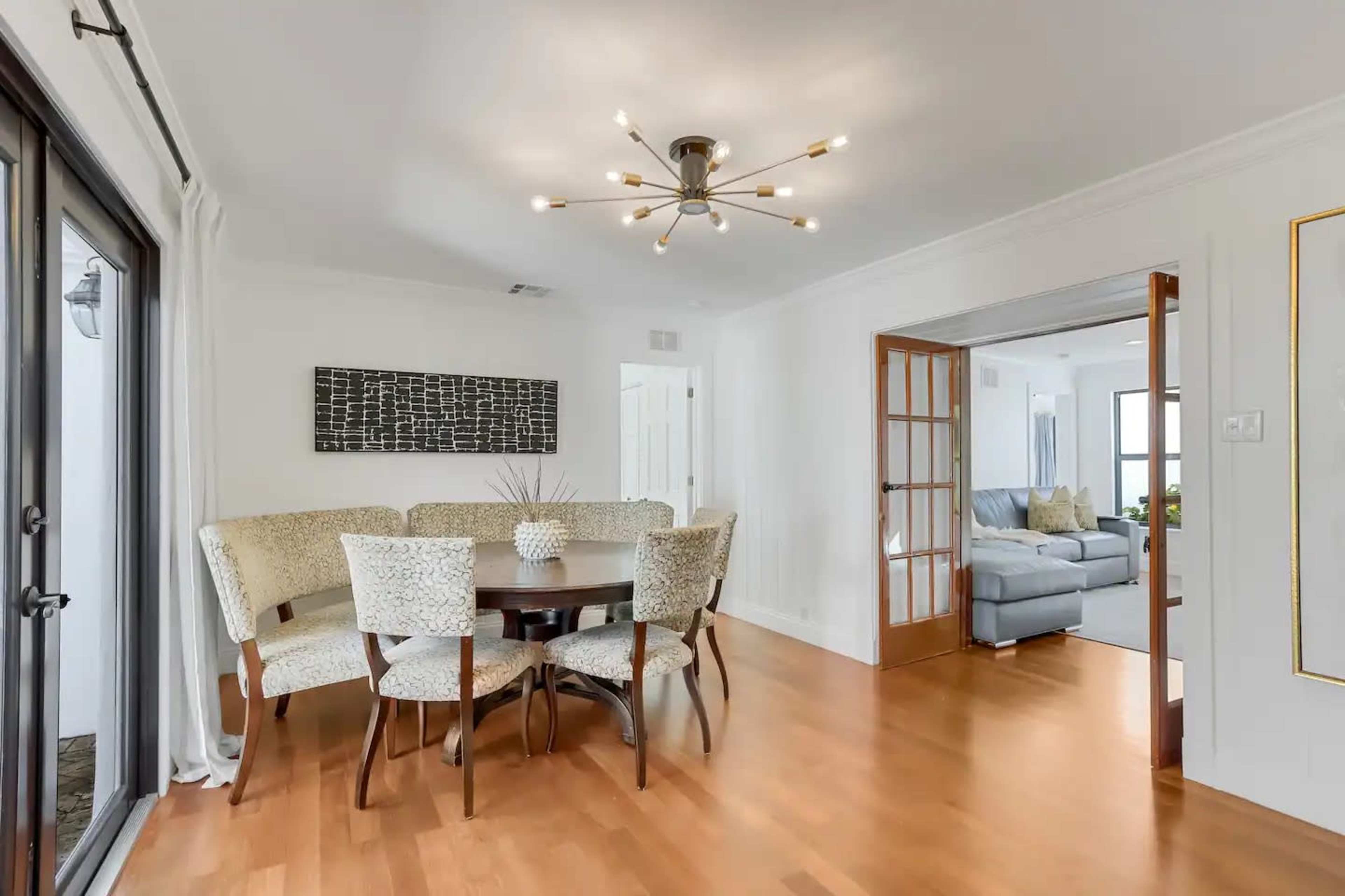 A modern dining area features a circular table surrounded by upholstered chairs, with a light fixture above and a view into a living space.