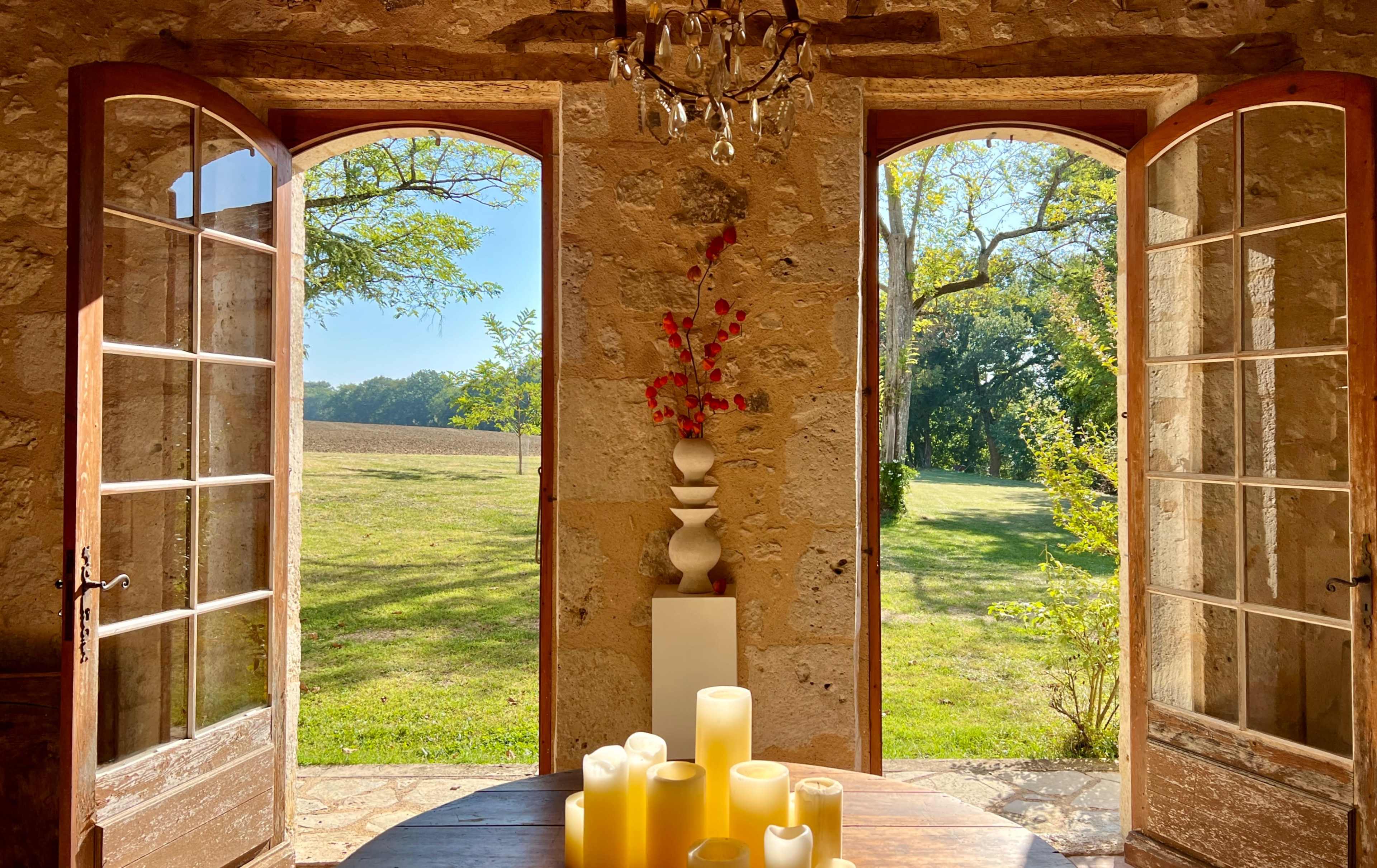A sunlit room features two open wooden doors that lead to a grassy field, with a table adorned with yellow candles in the foreground.