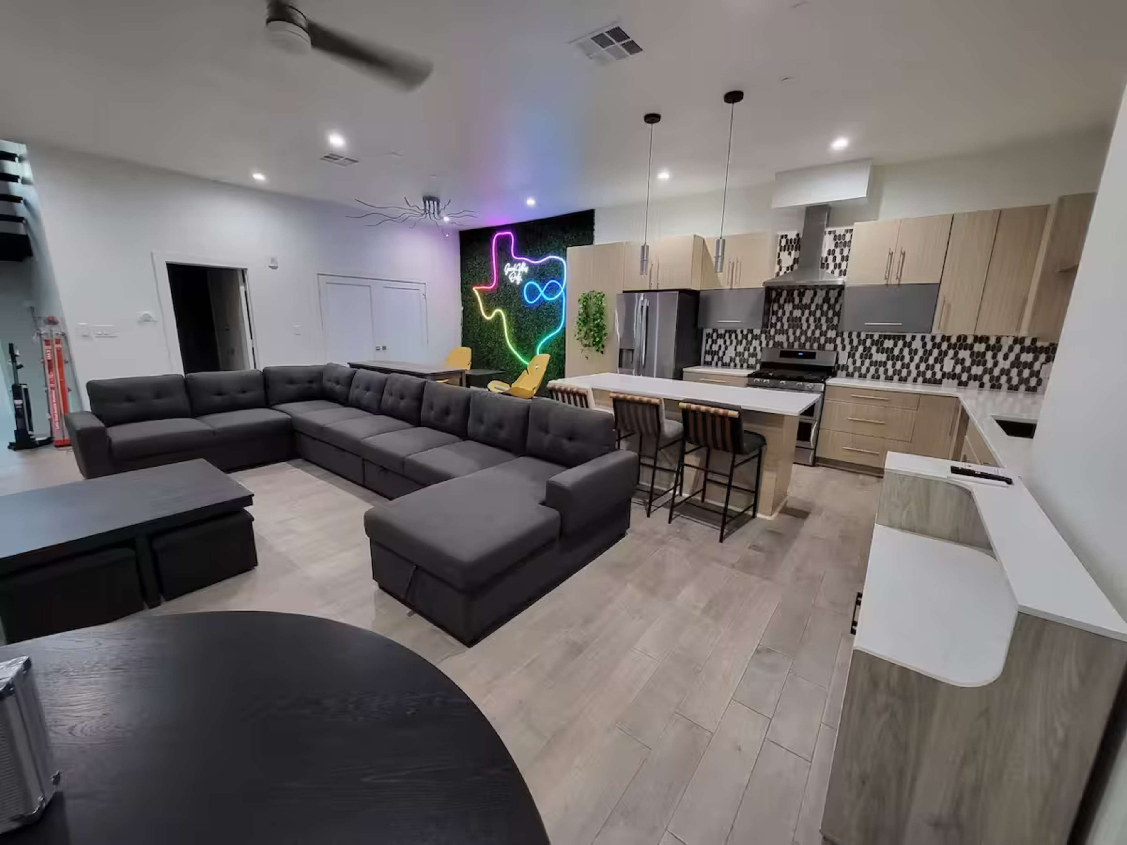 The image shows a modern living area with a large sectional sofa, a kitchen featuring light wood cabinets and a patterned backsplash, and a neon wall art piece in the background.