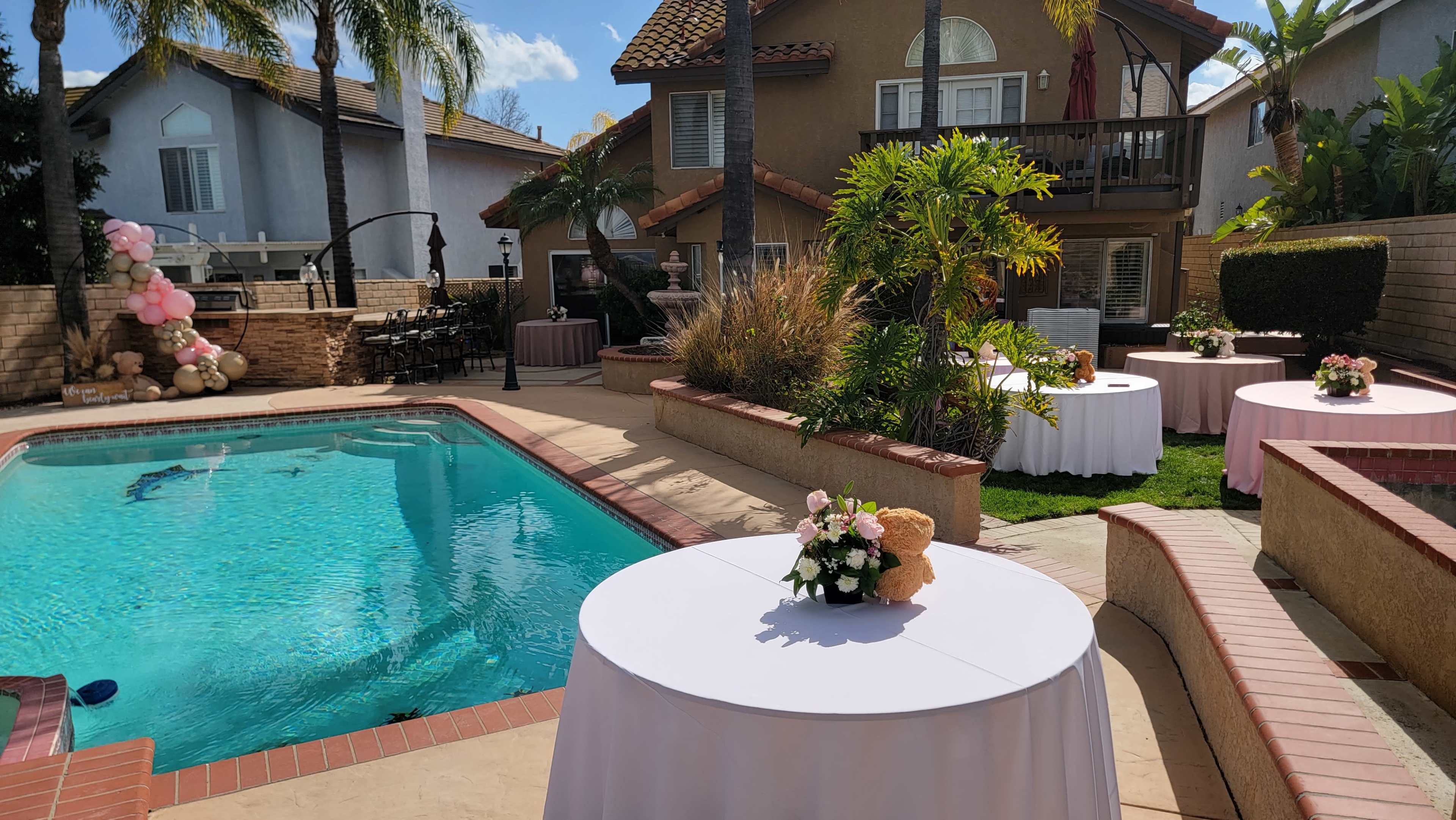 The scene shows a backyard with a swimming pool, several round tables set with white tablecloths, and decorative floral arrangements, surrounded by palm trees and a house in the background.