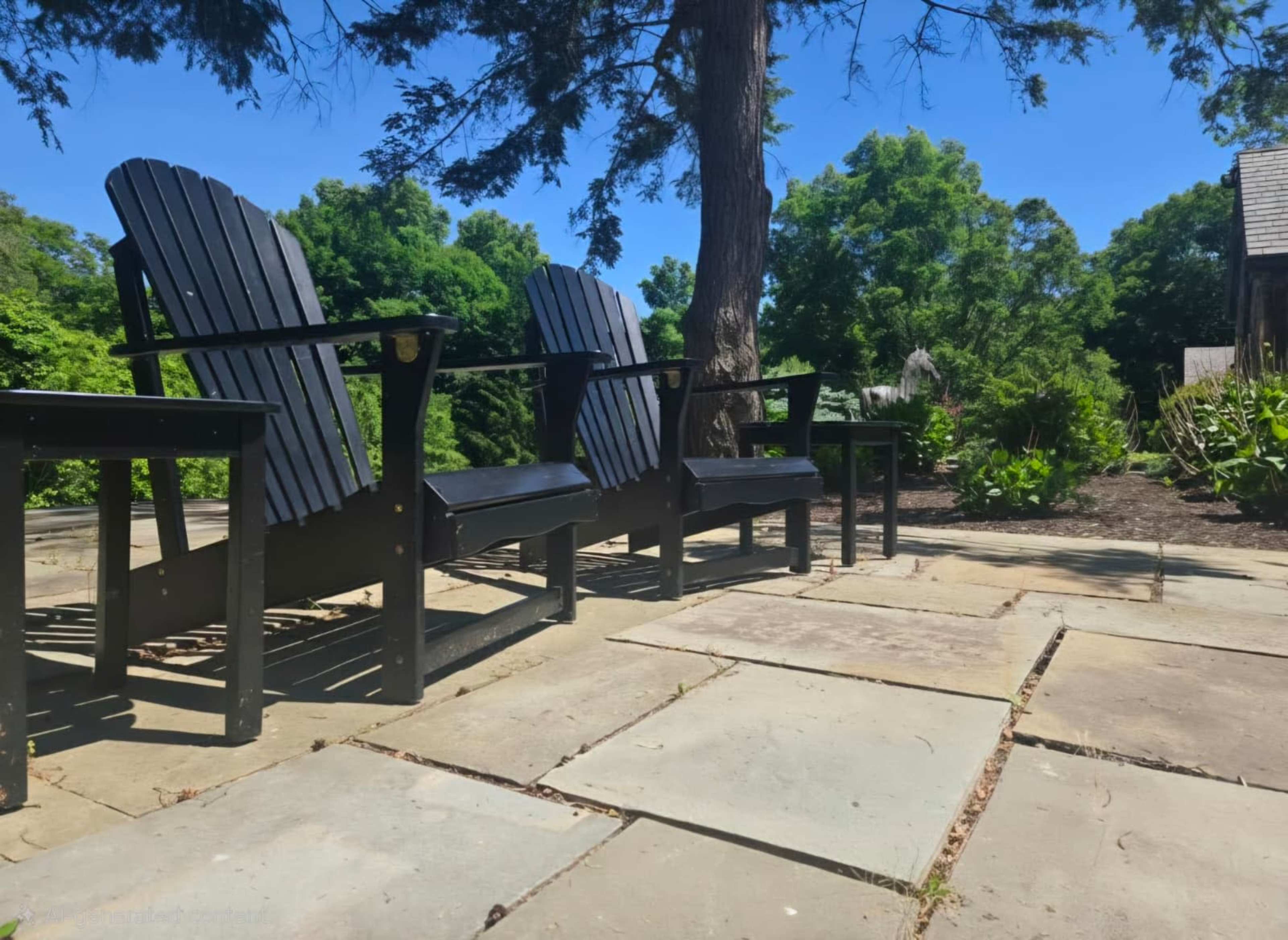 Two black Adirondack chairs sit on a stone patio surrounded by lush green foliage and trees.
