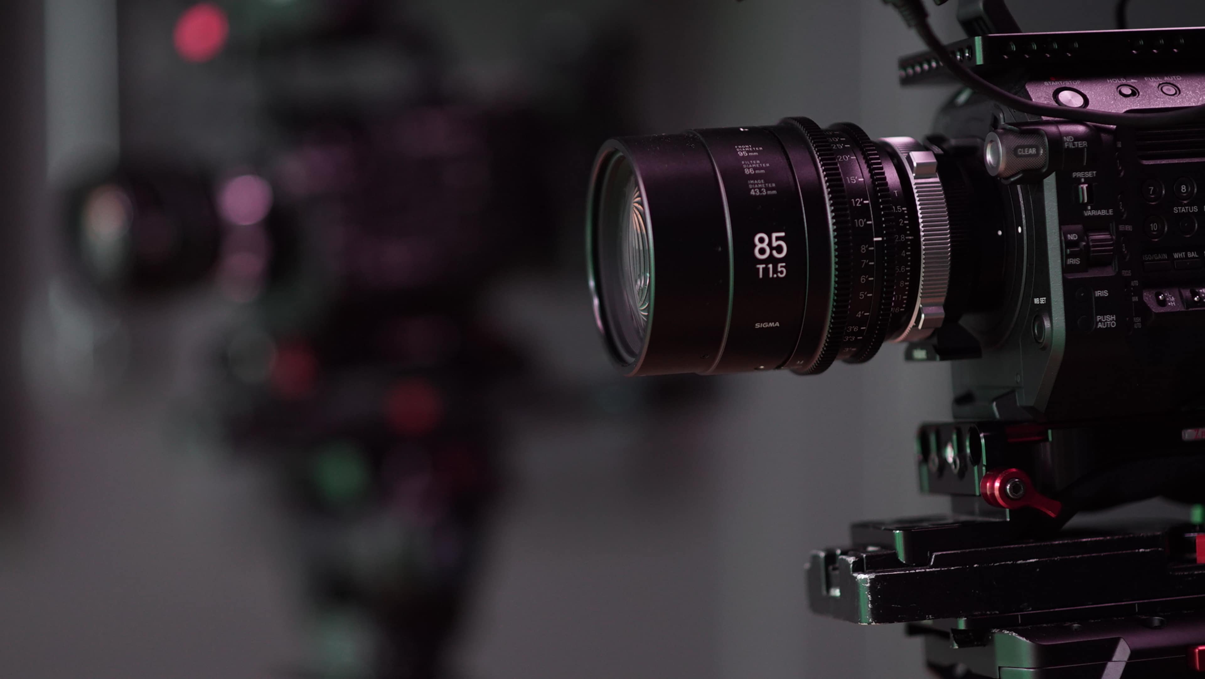 A close-up of a camera lens labeled "85 T1.5" mounted on a professional camera, with another camera slightly blurred in the background.