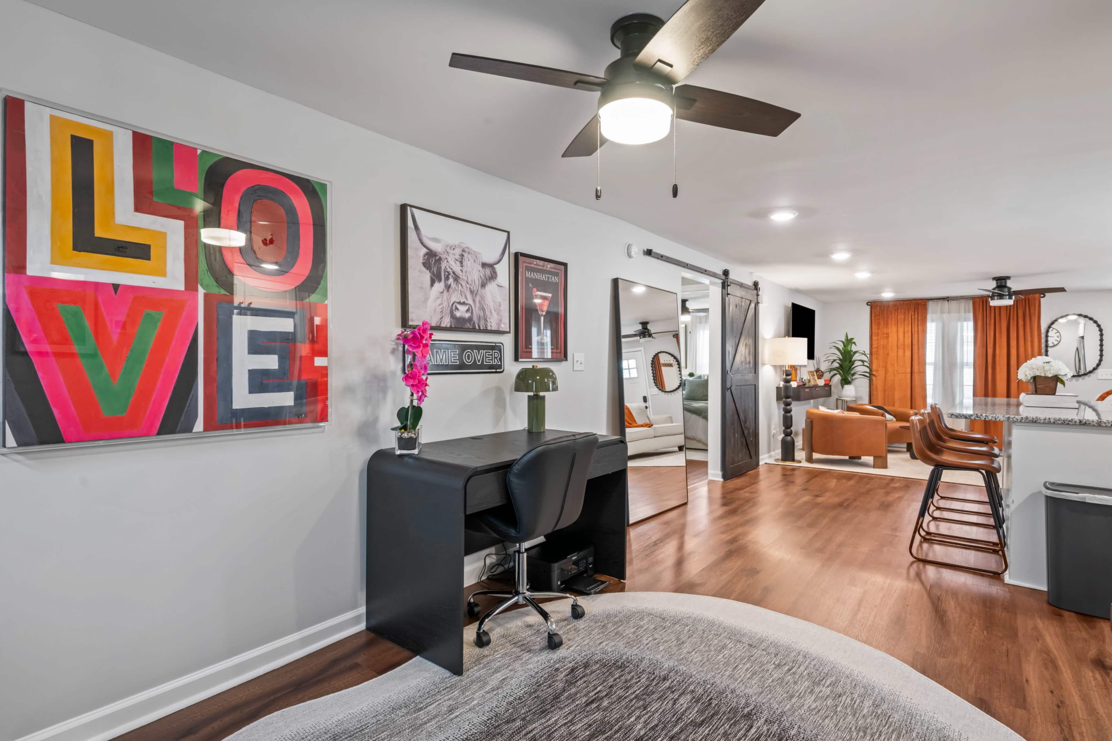 The image shows a modern living space featuring a desk with a chair, colorful wall art, and a comfortable seating area with orange armchairs and a visible kitchen.