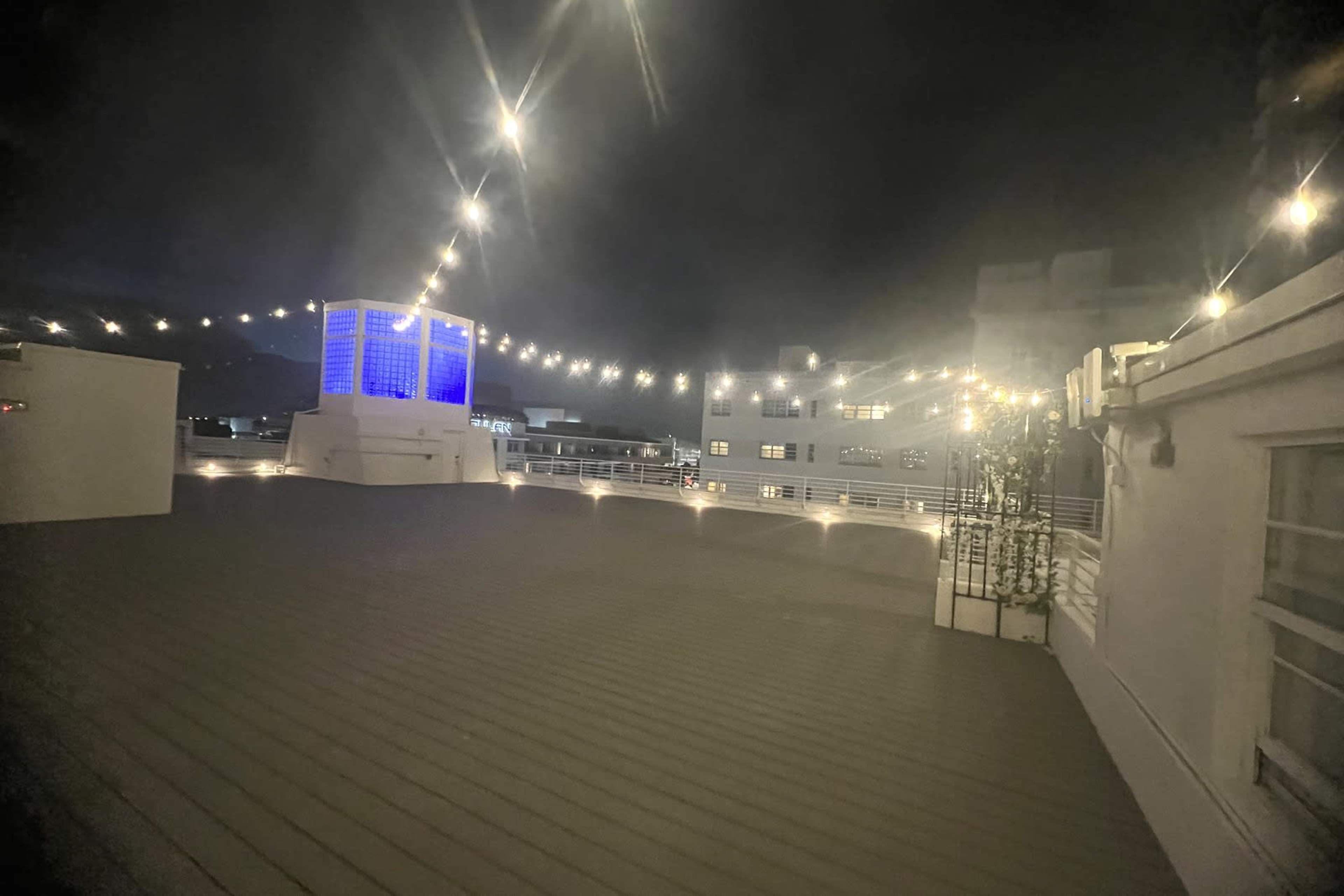 The image shows a rooftop at night, illuminated by string lights and featuring a blue illuminated structure in the background.