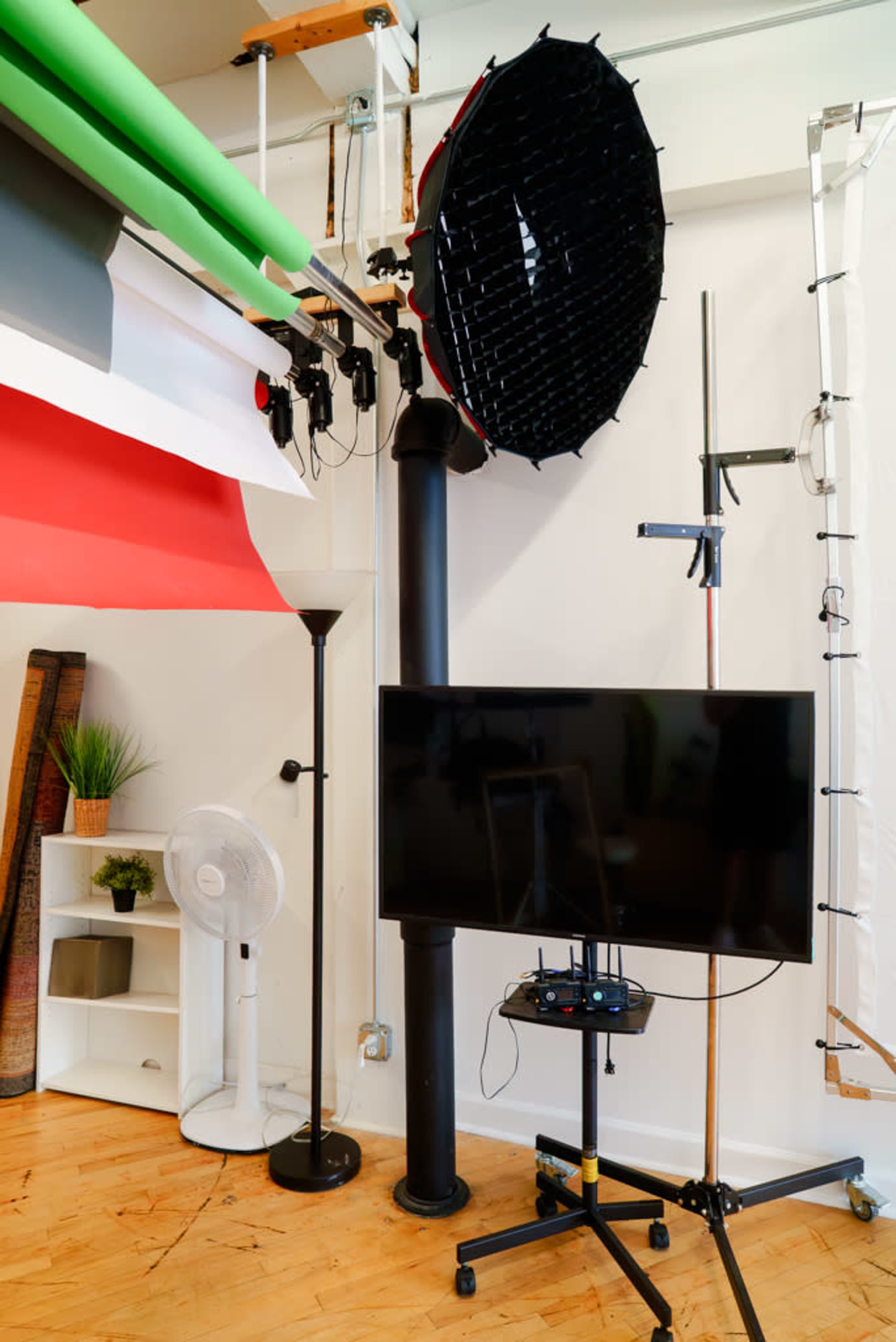 The image shows a photography studio setup featuring a large softbox light, a black television on a stand, and a few studio equipment pieces, including a fan and a light pole.