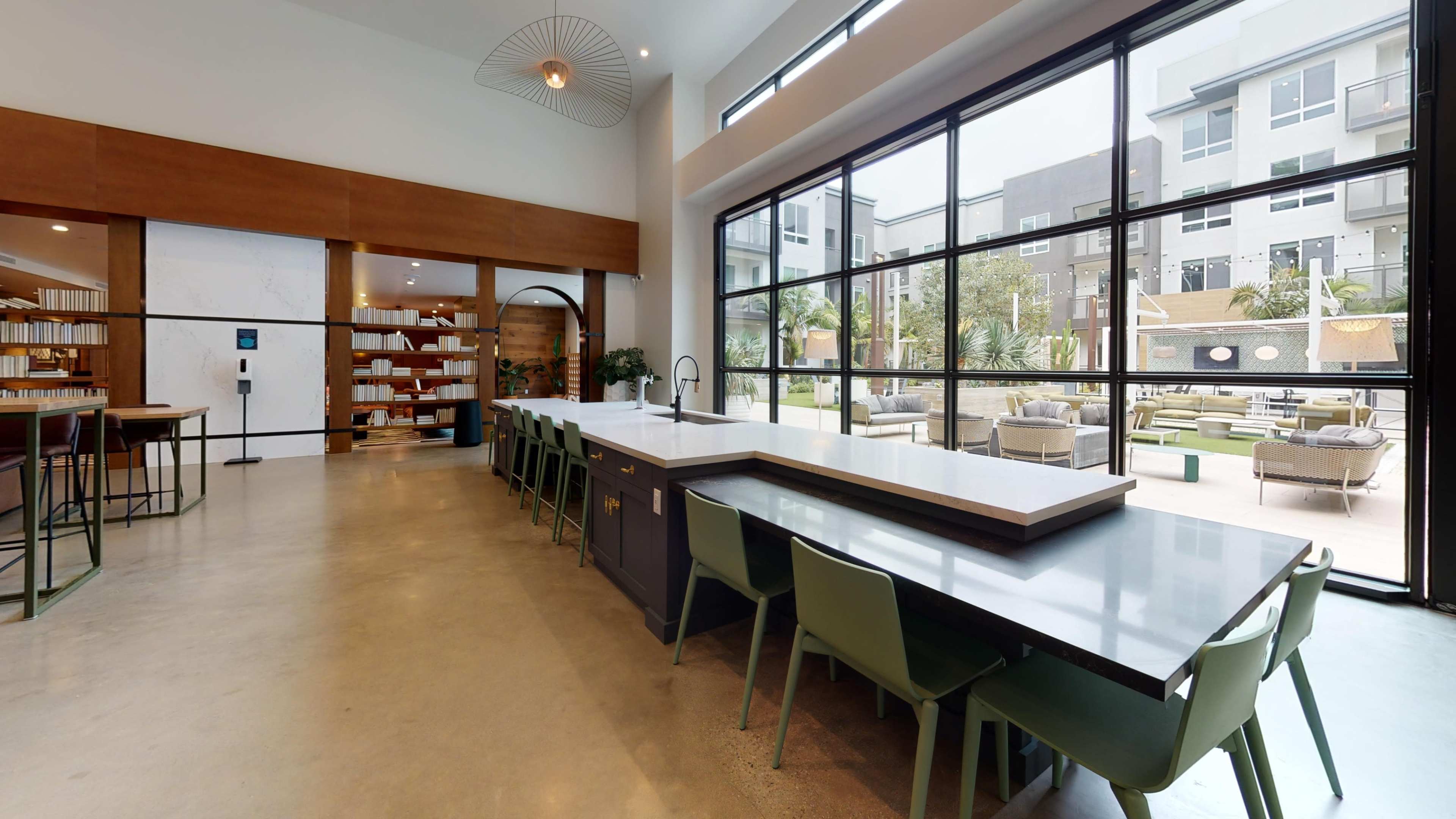 A modern kitchen area features a long countertop with seating and large windows overlooking an outdoor patio with lounge furniture.