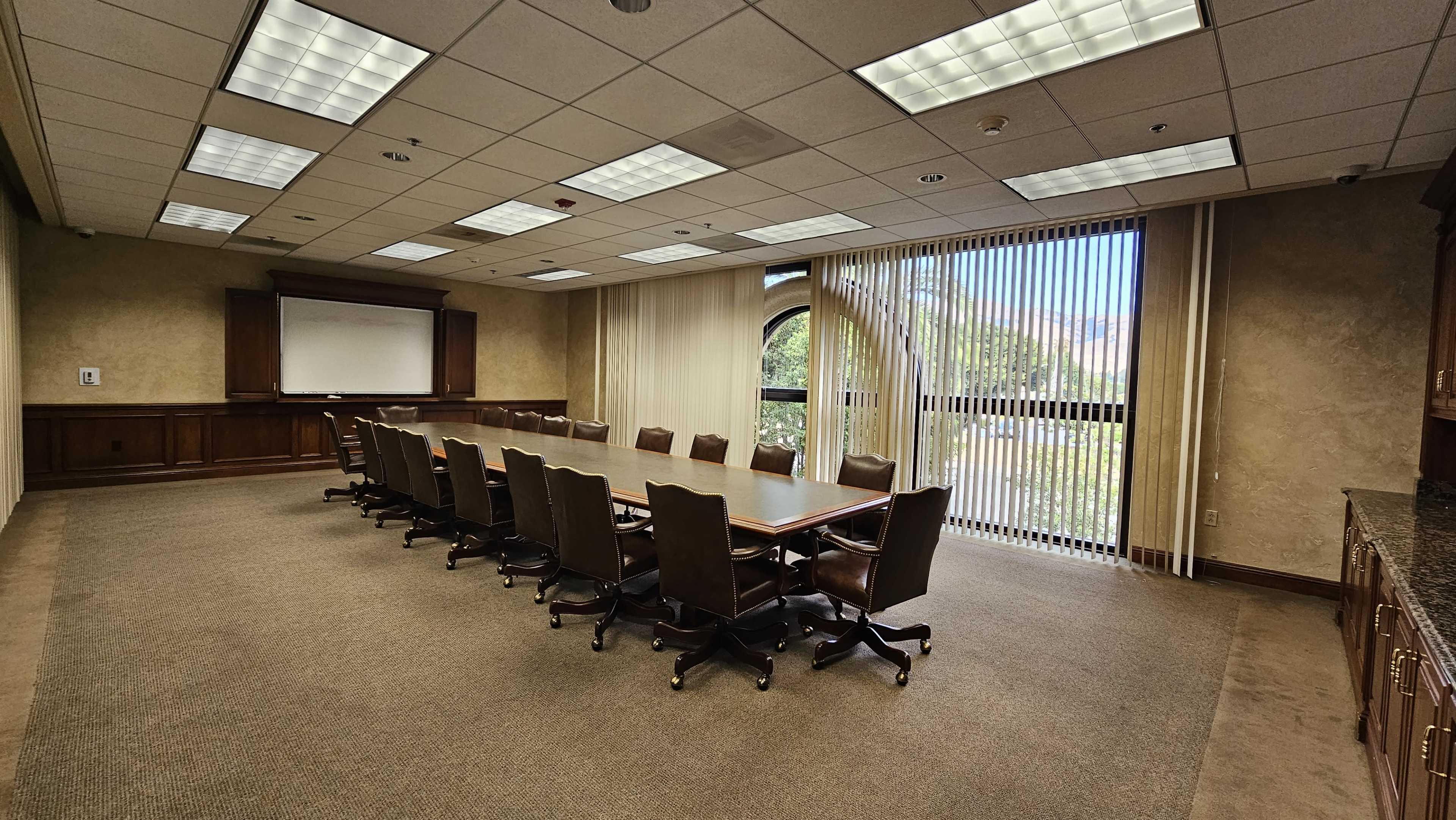 A large, empty conference room features a long wooden table surrounded by black upholstered chairs, with large windows letting in natural light and vertical blinds drawn to the side.