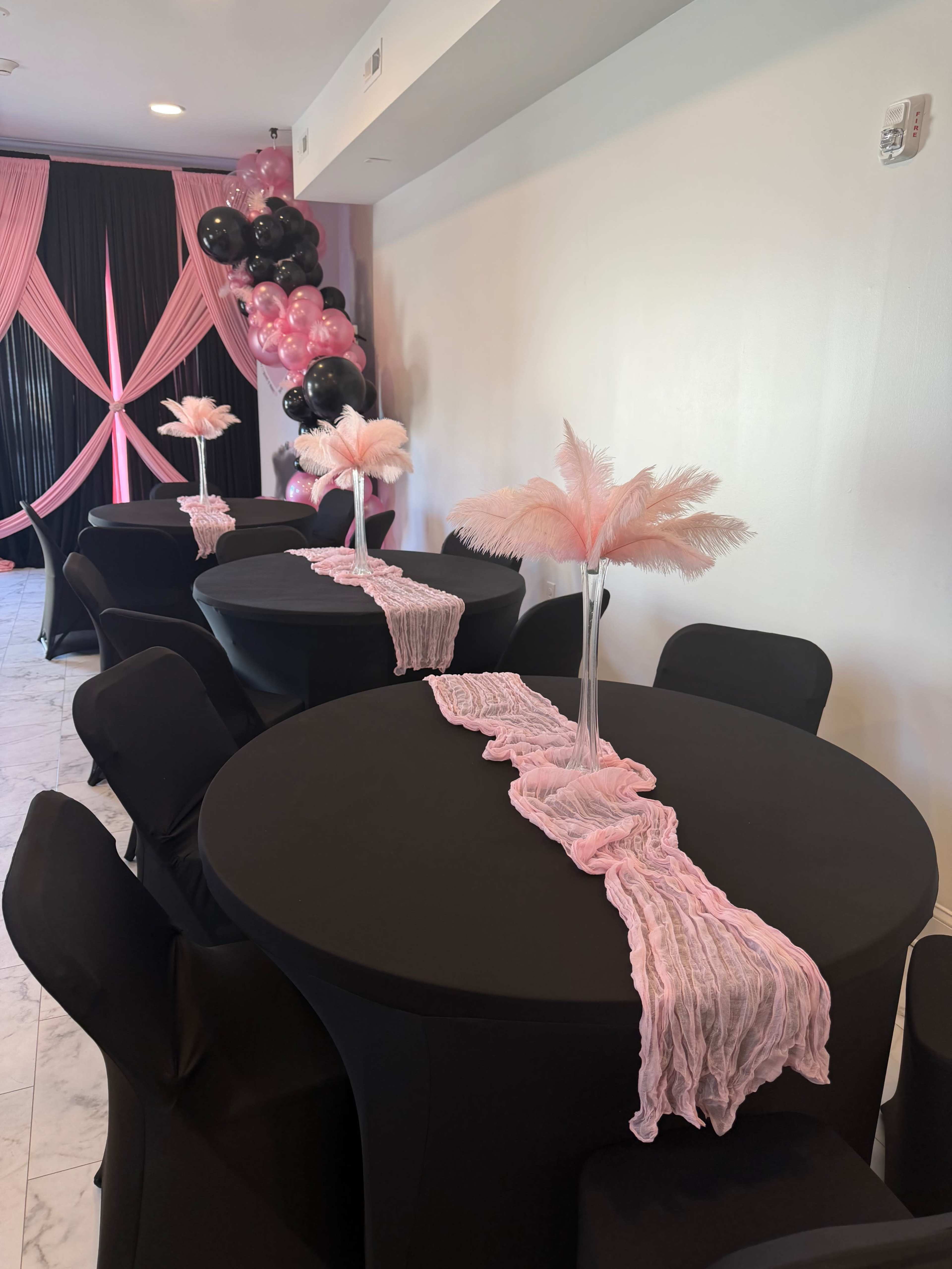 The image shows a banquet setup with black circular tables adorned with pink accent decorations and tall feather centerpieces, set against a backdrop of black and pink balloons.