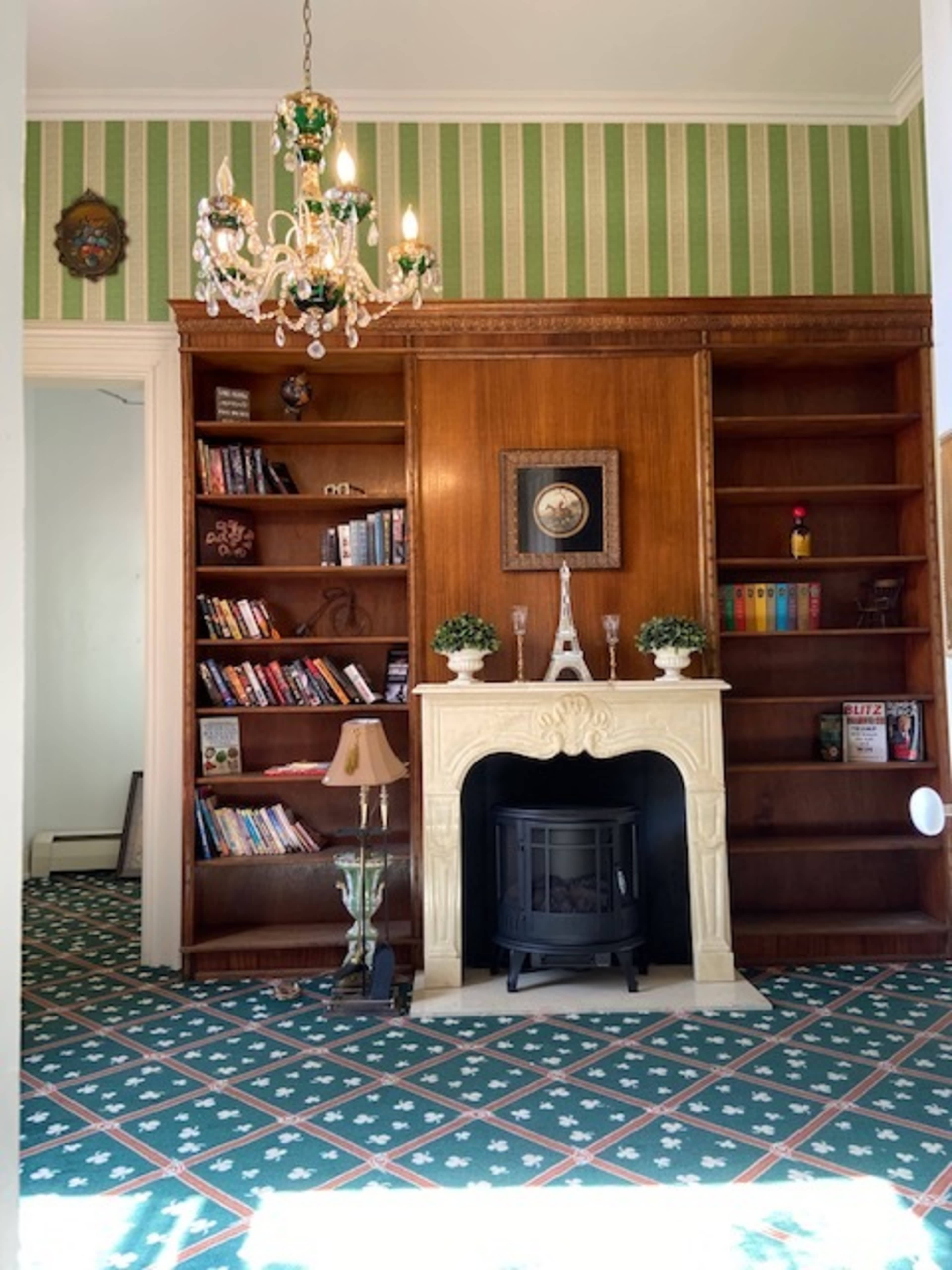 A cozy room featuring a wooden bookshelf filled with books, a decorative fireplace with an ornate mantel, and a chandelier hanging from the ceiling.