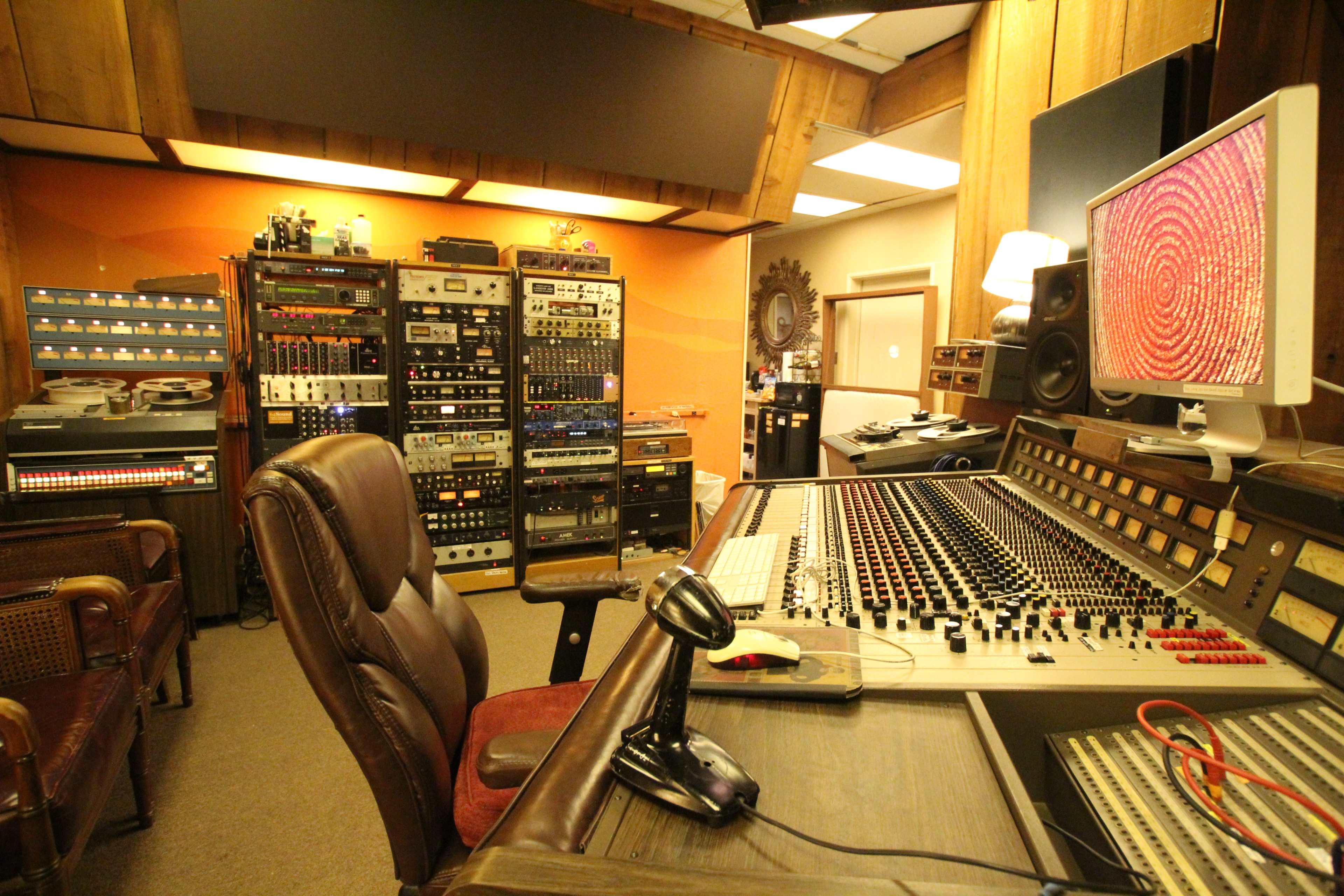 A recording studio with a large mixing console, various audio equipment racks, and a leather office chair.