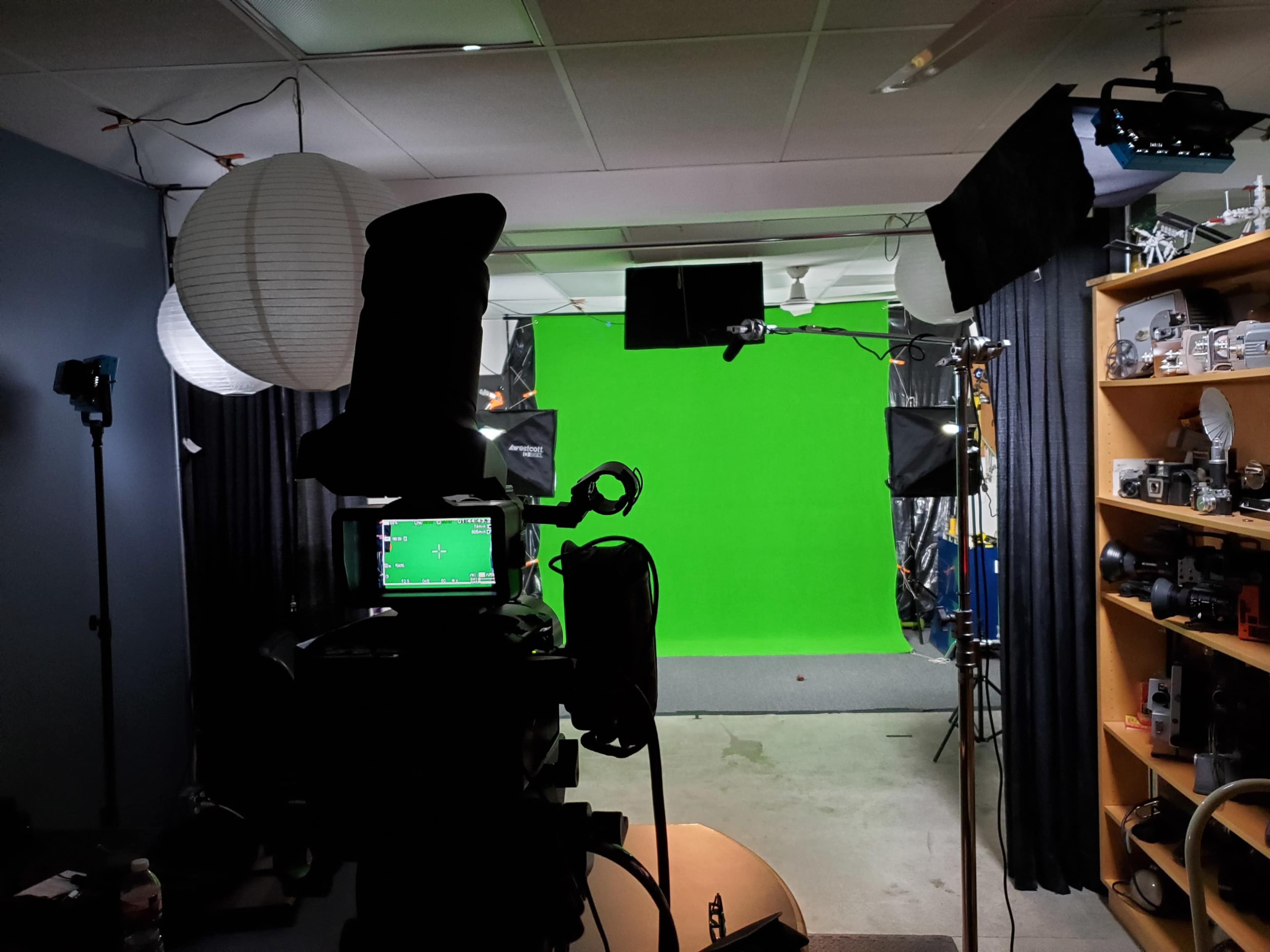 A studio setup with a green screen backdrop, various lighting equipment, and a camera positioned in front of a cluttered shelf containing photographic gear.