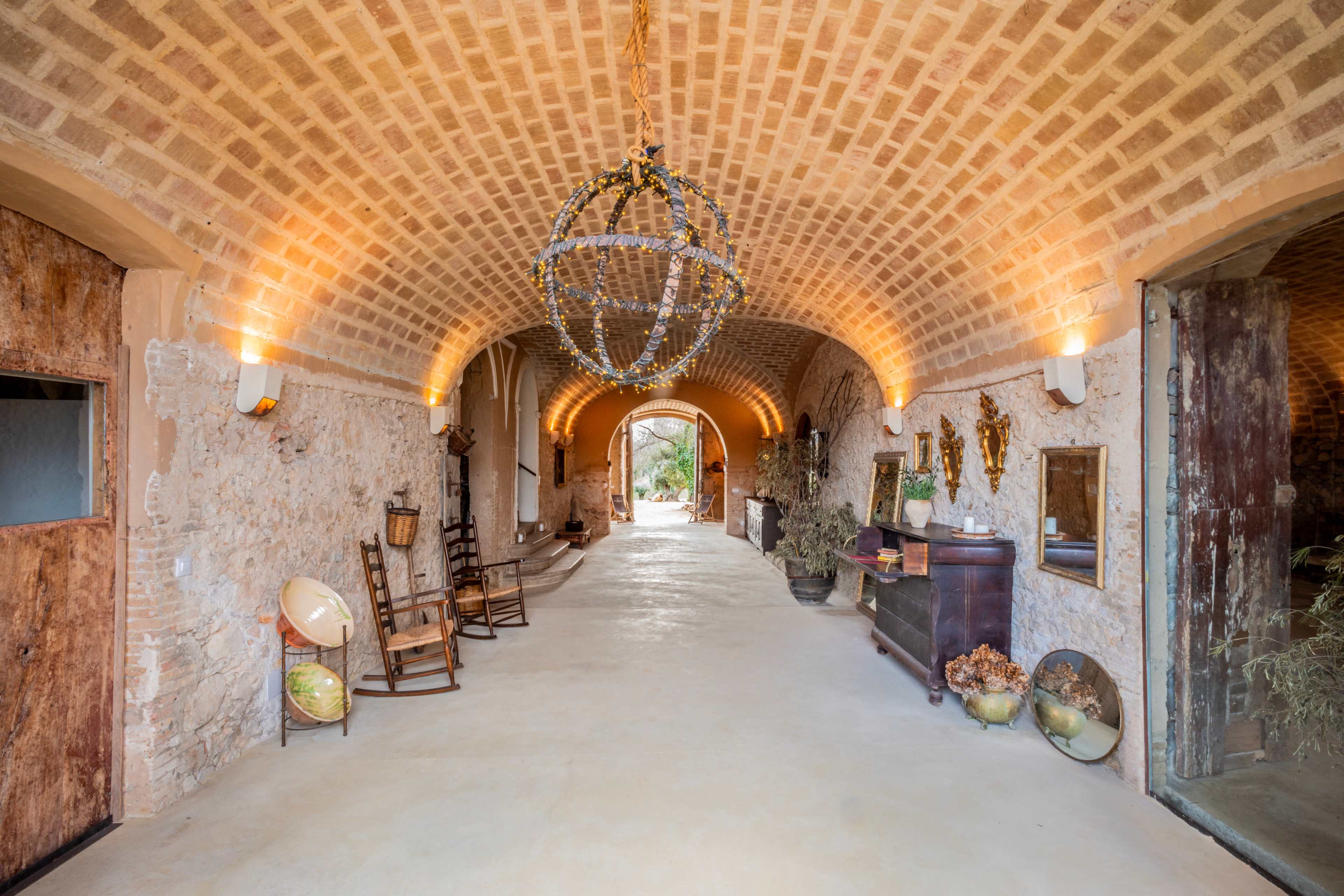 The image shows a spacious, arched hallway with textured stone walls, wooden doors, and decorative elements, leading to an outdoor view.