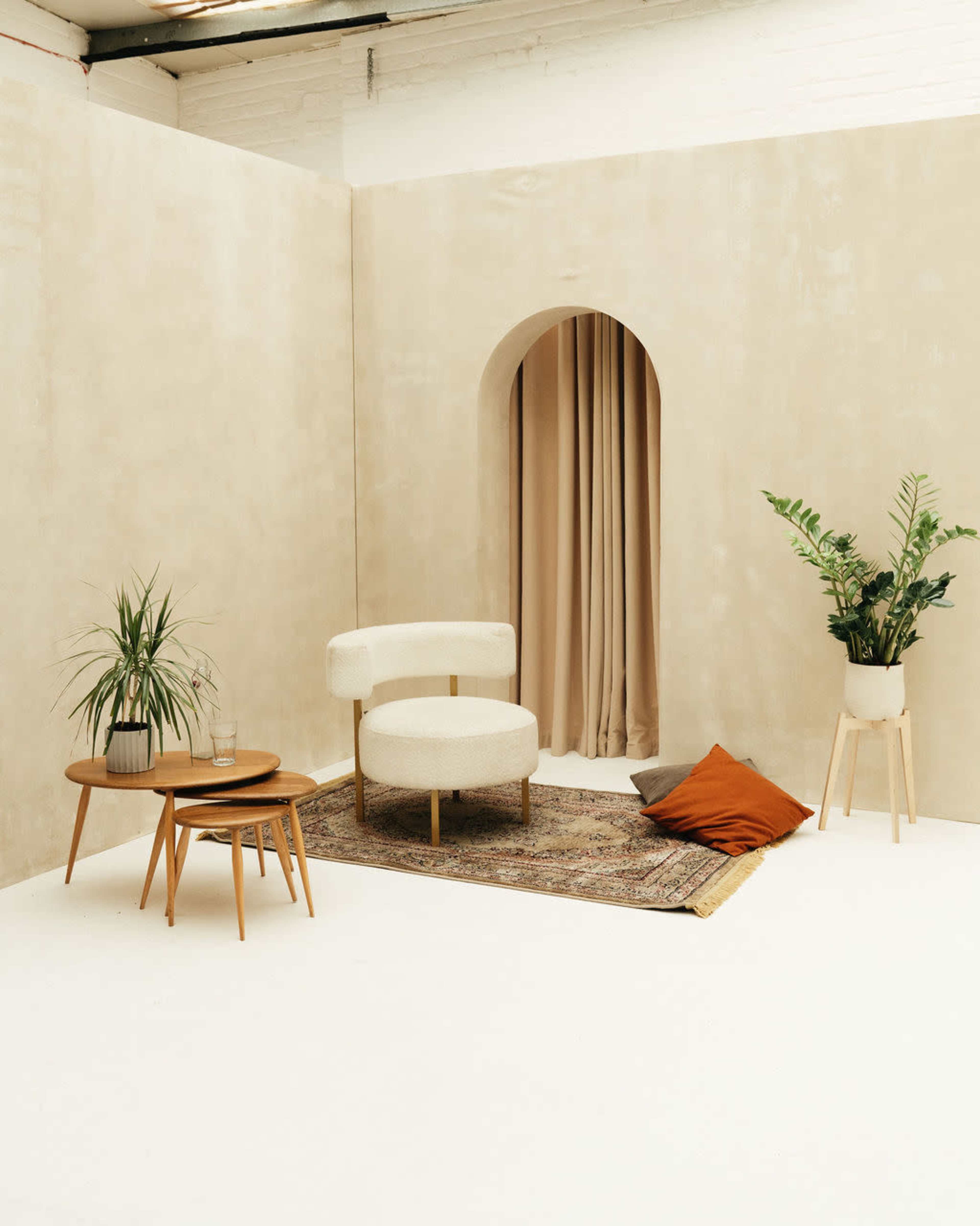 The image shows a minimalist interior space with a curved sofa, a round coffee table, a decorative rug, and plants arranged against a neutral-colored wall.