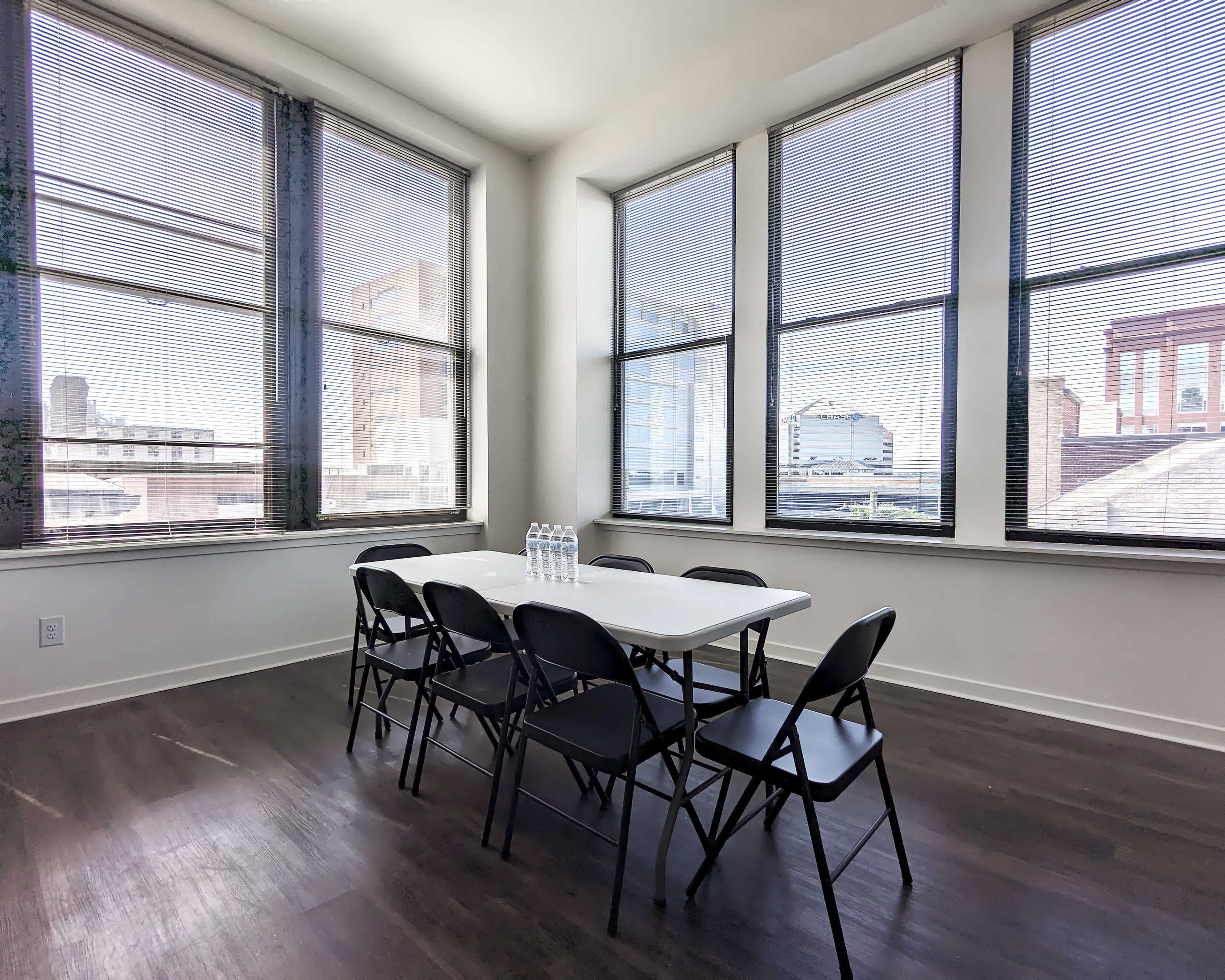 A conference room with a white table and six black chairs is illuminated by natural light from large windows.