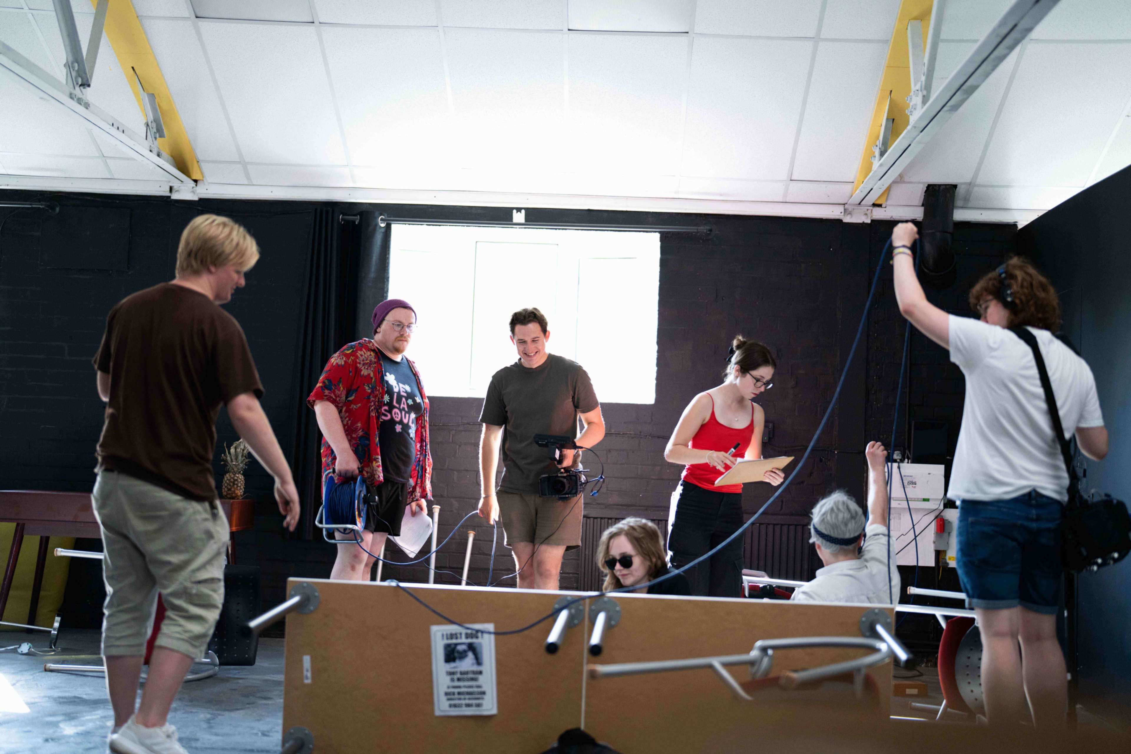 A group of eight people is engaged in various activities inside a studio space, with a large window providing natural light.