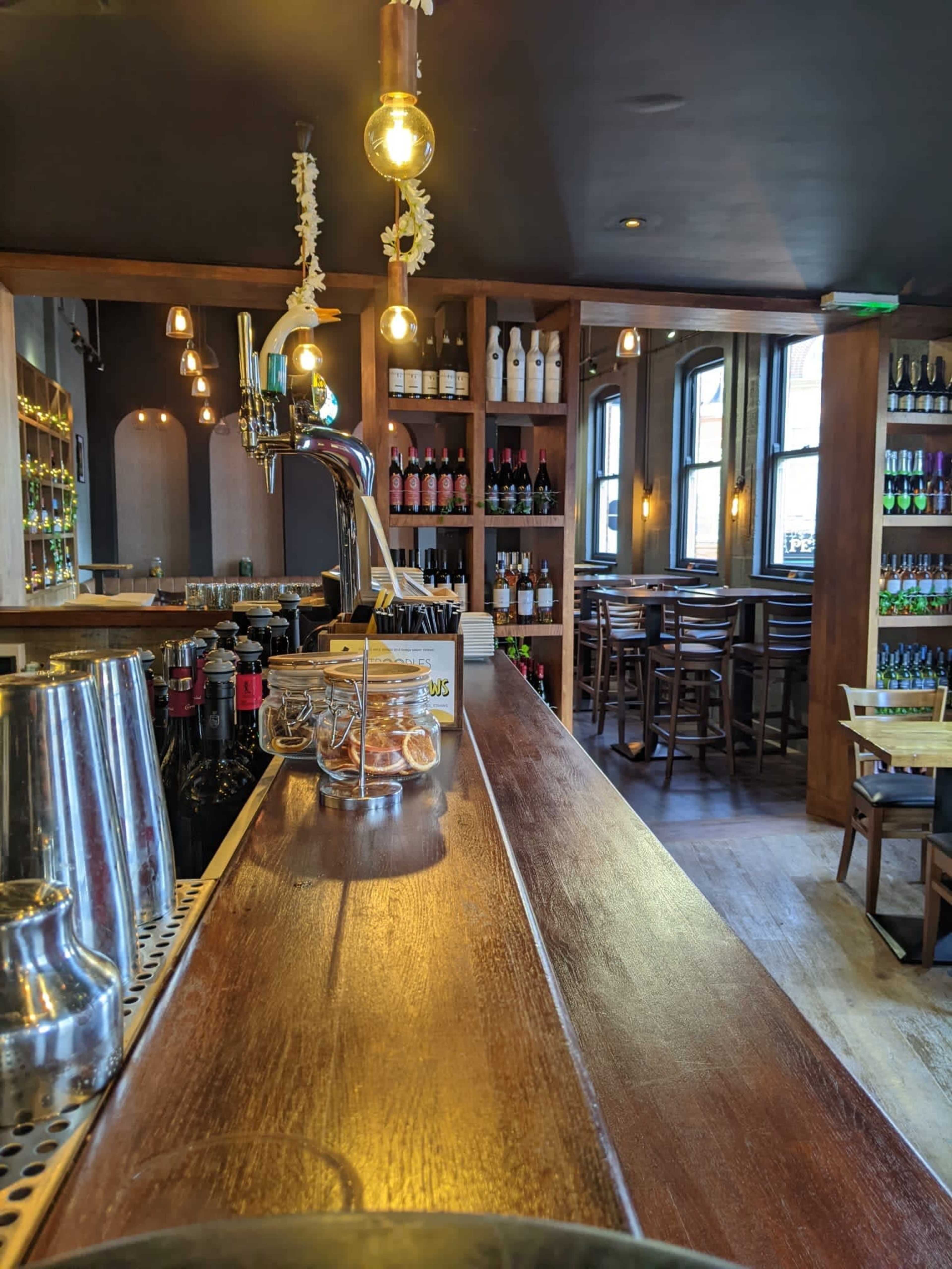 A wooden bar with a beer tap and shelves filled with bottles of wine and beer, set in a cozy interior with bar stools and warm lighting.