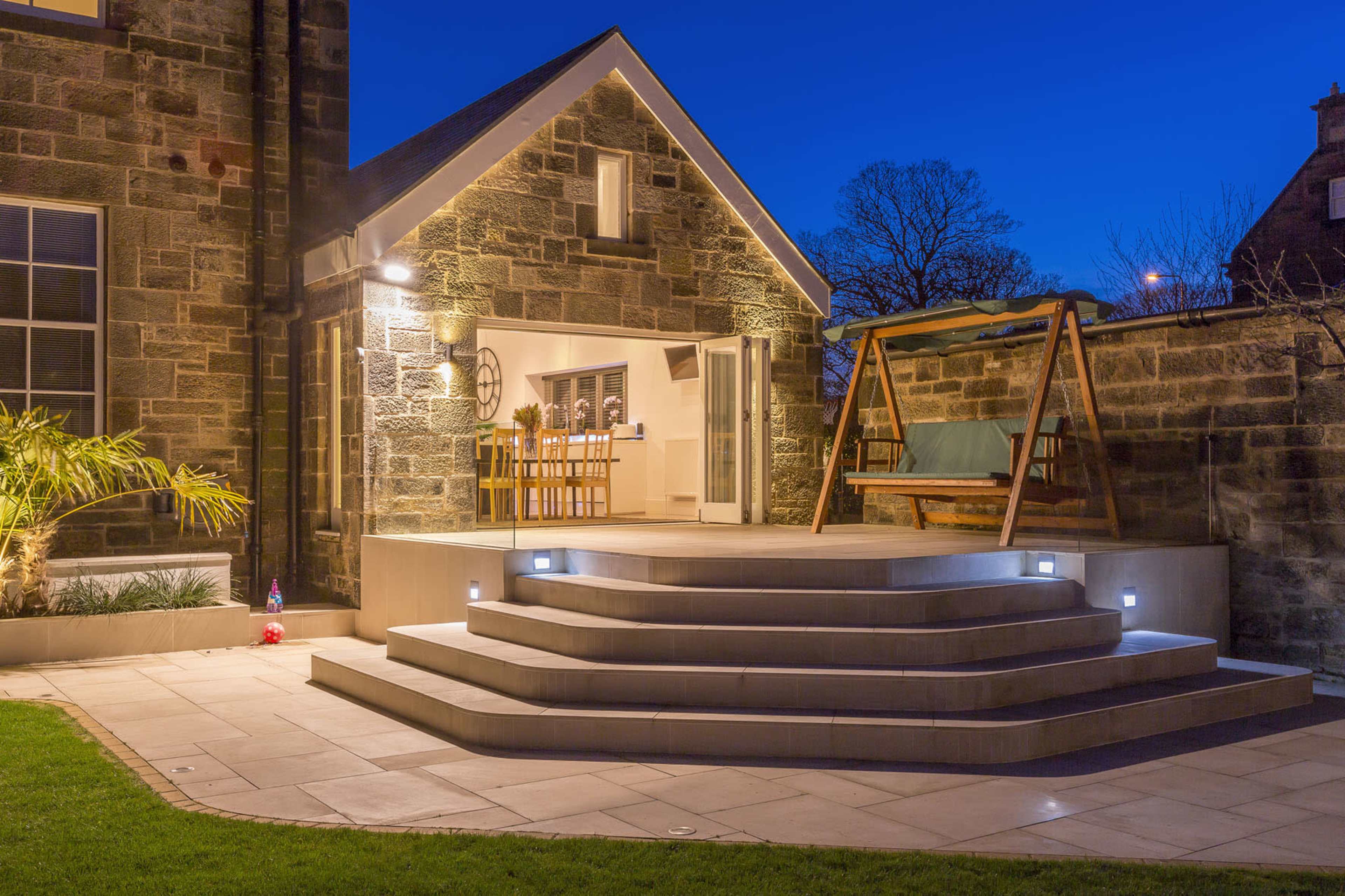 A modern stone house features a tiered patio with outdoor seating, surrounded by a well-maintained lawn, illuminated under a twilight sky.