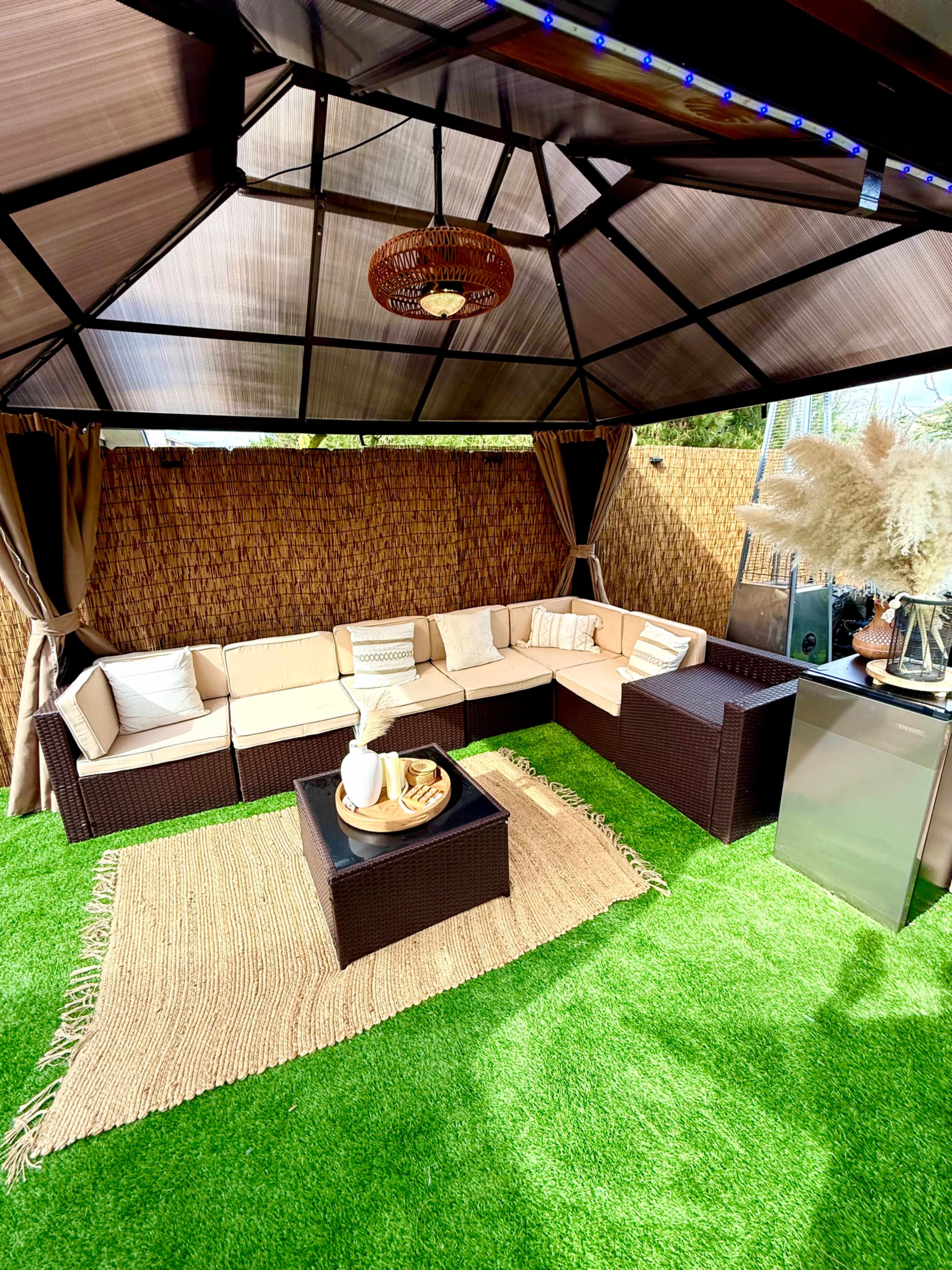 The image shows a cozy outdoor seating area with a sectional sofa, a coffee table, and artificial grass flooring under a gazebo.