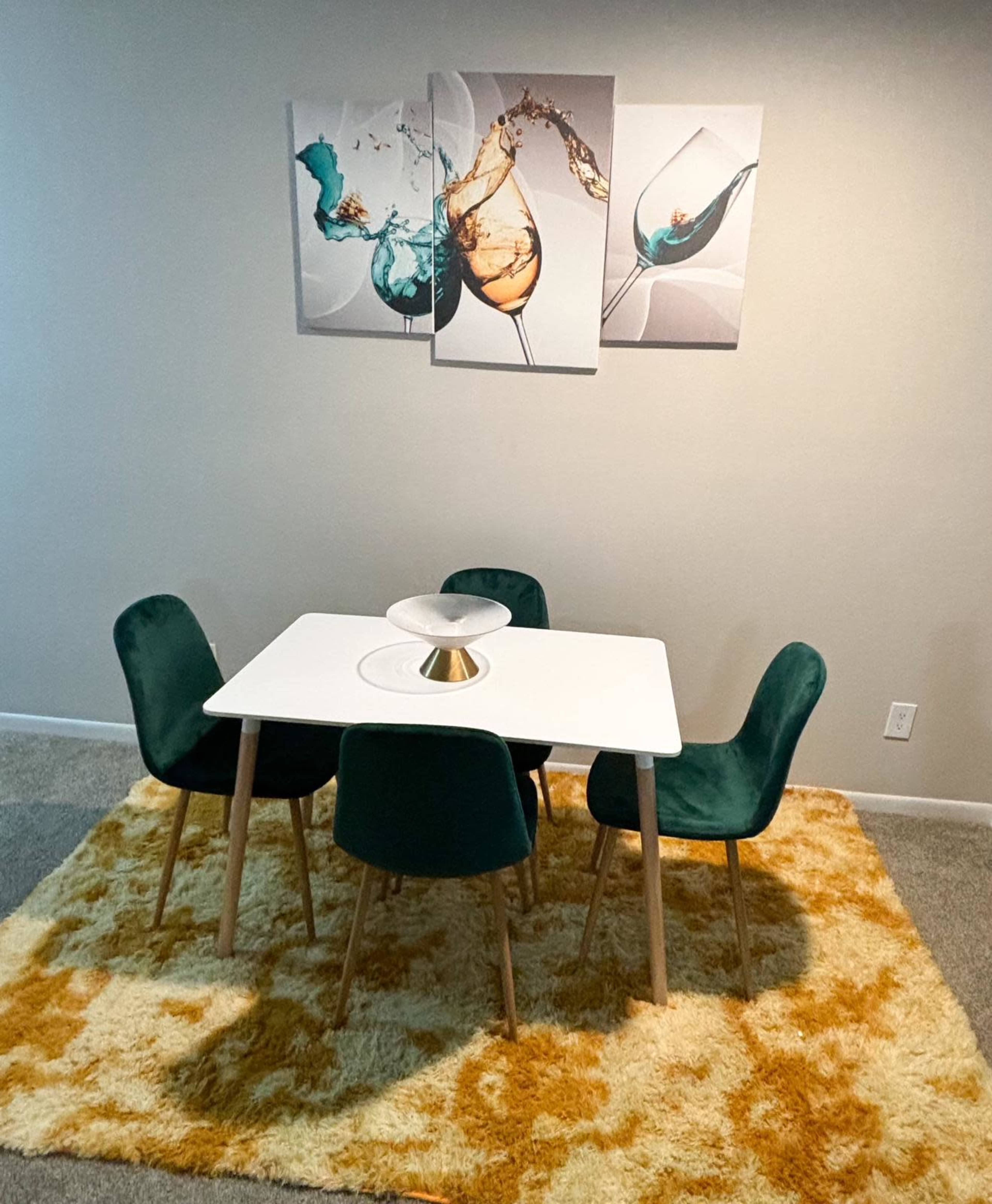 A modern dining area features a square white table surrounded by four green chairs on a large, yellow and cream rug, with abstract art depicting glasses of beverages on the wall.