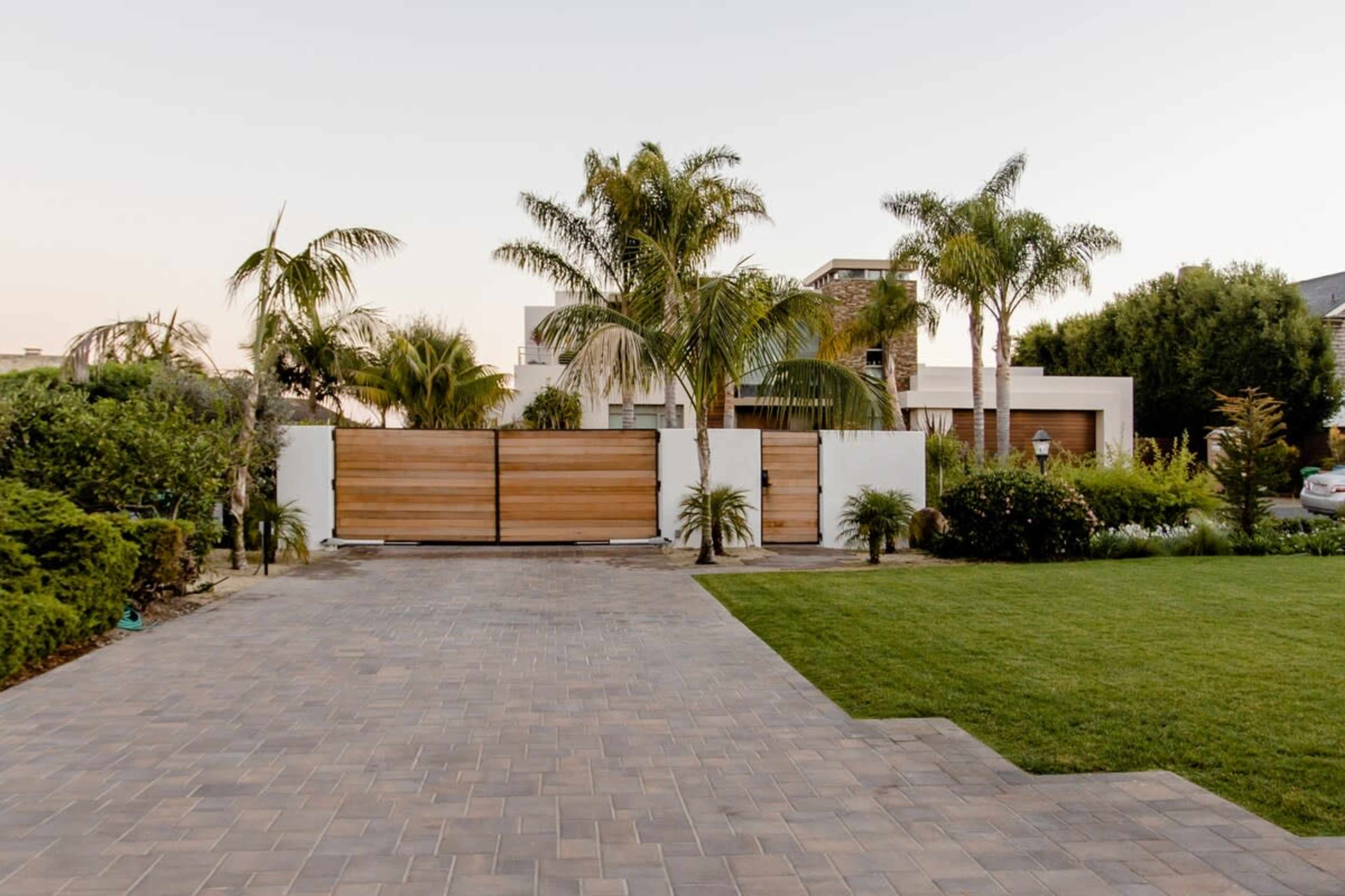 A modern house entrance with wooden gates surrounded by palm trees and a paved driveway leading to a lawn.