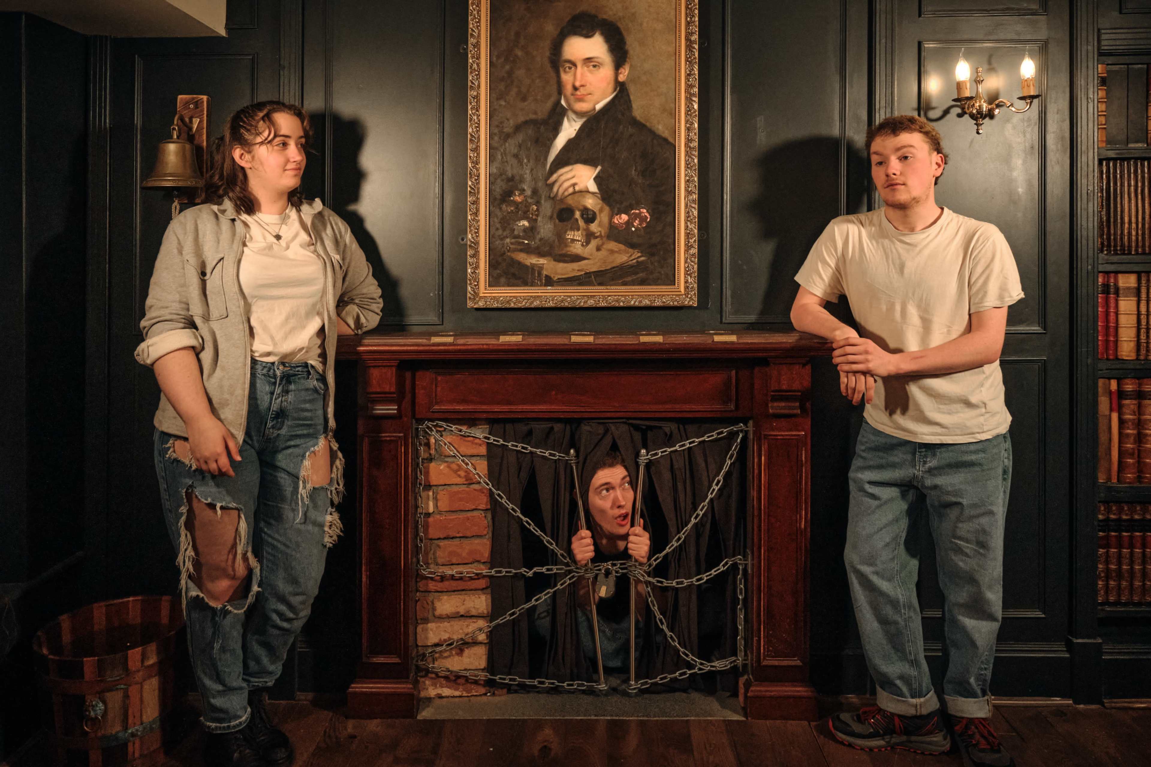 Two people stand beside a fireplace framed by a portrait, with one person appearing to peek out from behind a set of chains draped across the fireplace opening.