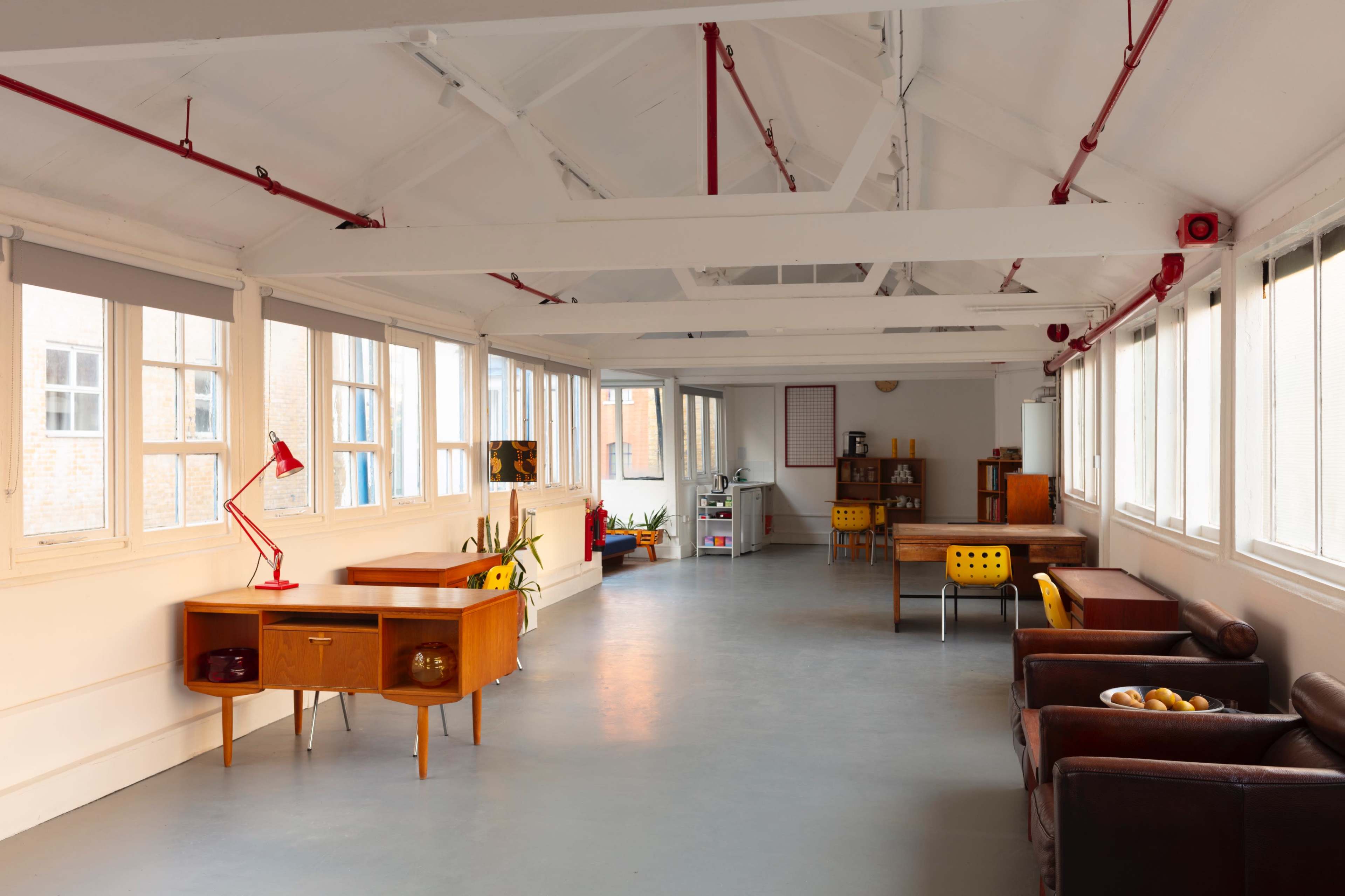 The interior of a bright, open space features wooden furniture, including desks and chairs, with large windows allowing natural light to enter.