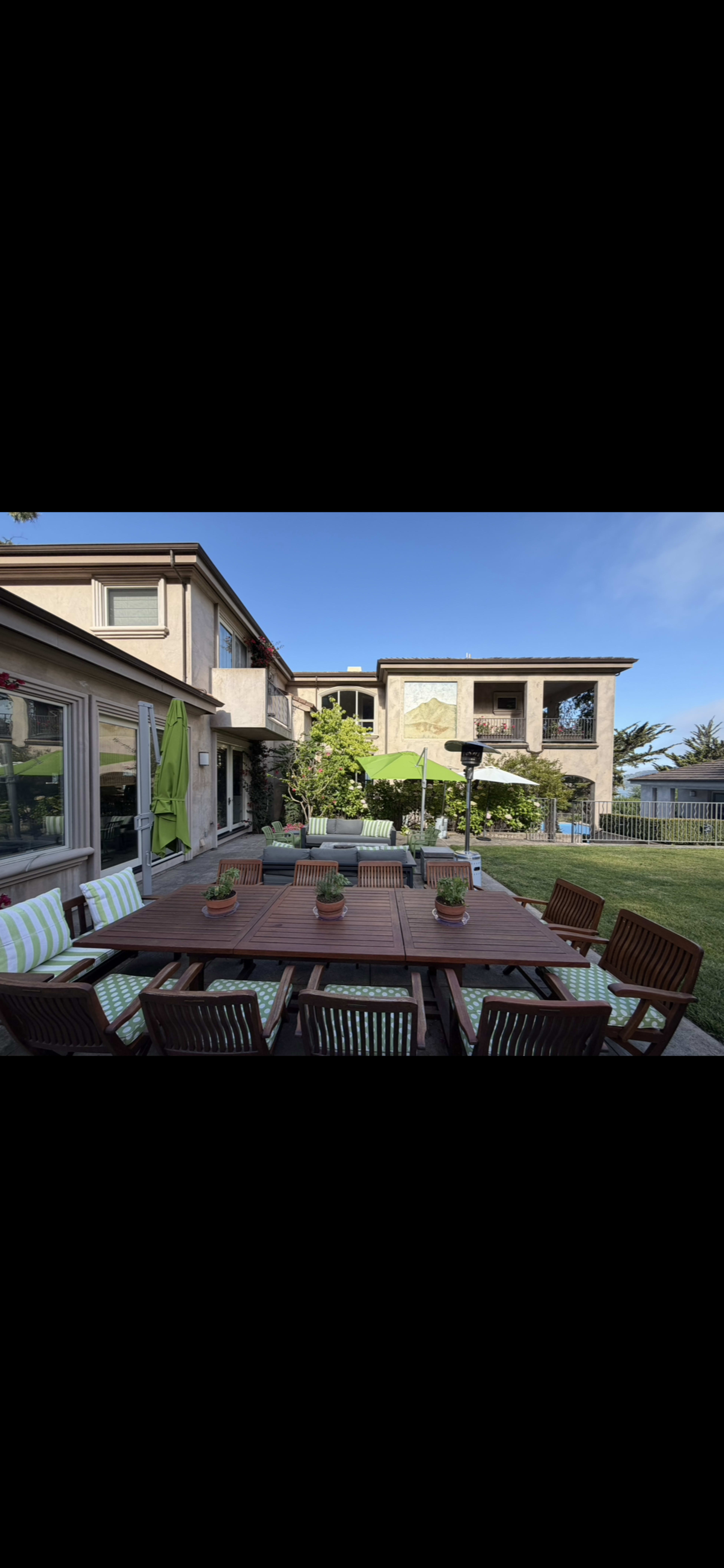 The image shows a spacious patio with a wooden dining table surrounded by chairs, overlooking a well-maintained garden and a two-story house.