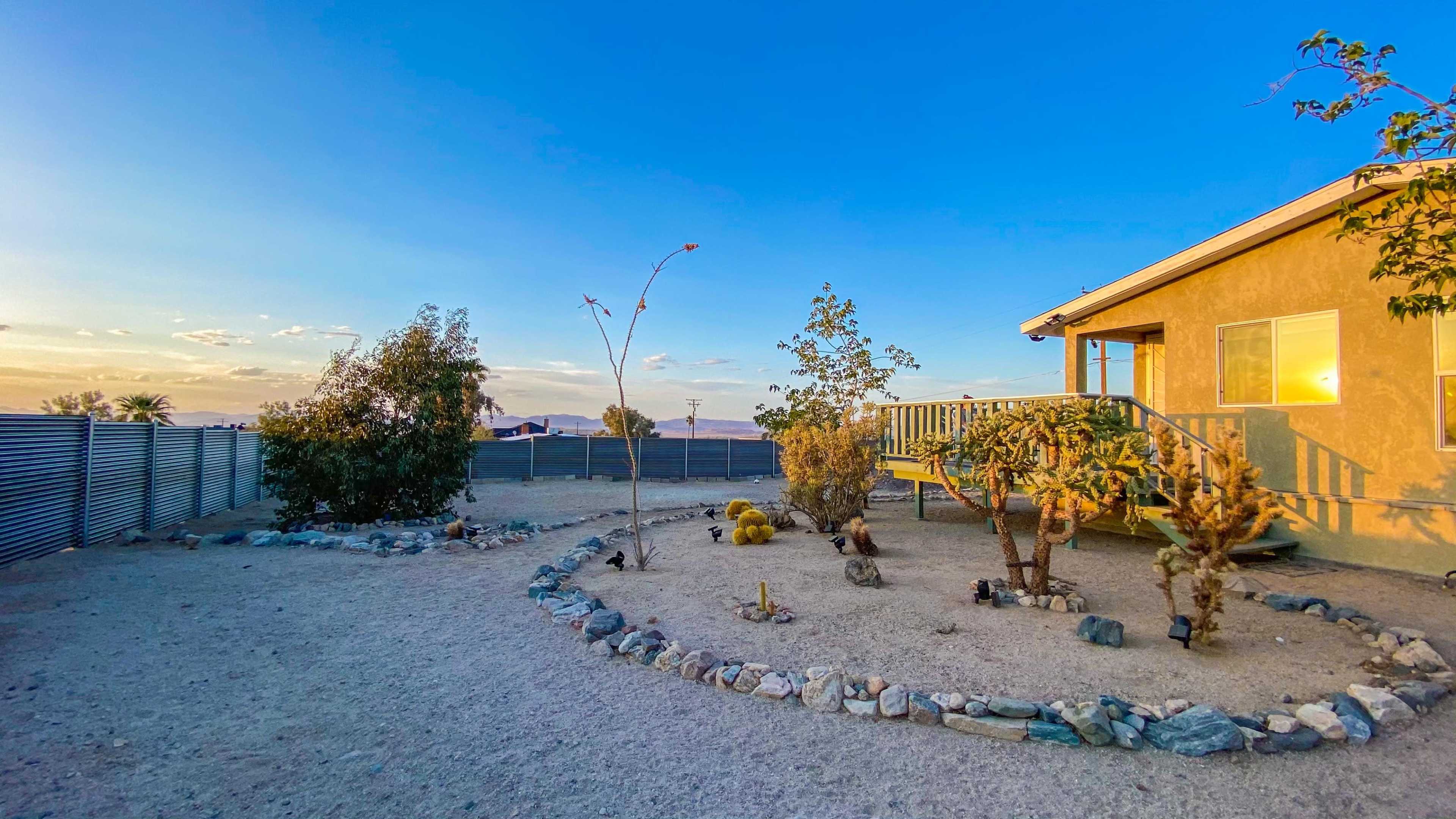 A house with a porch is surrounded by a gravel yard featuring several small trees and shrubs, with a circular rock border.