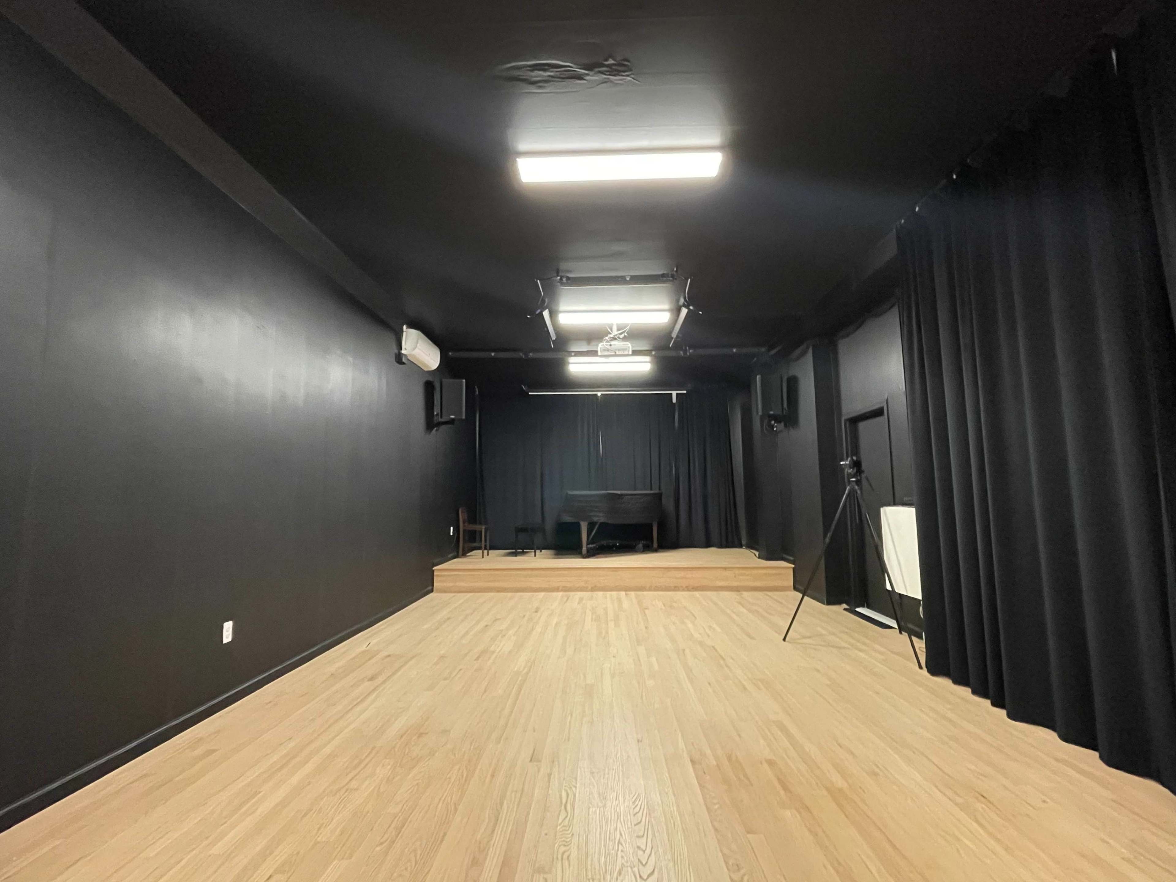 The image shows a dimly lit black box theater with wooden flooring, a small raised stage at the back, and black curtains lining the walls.