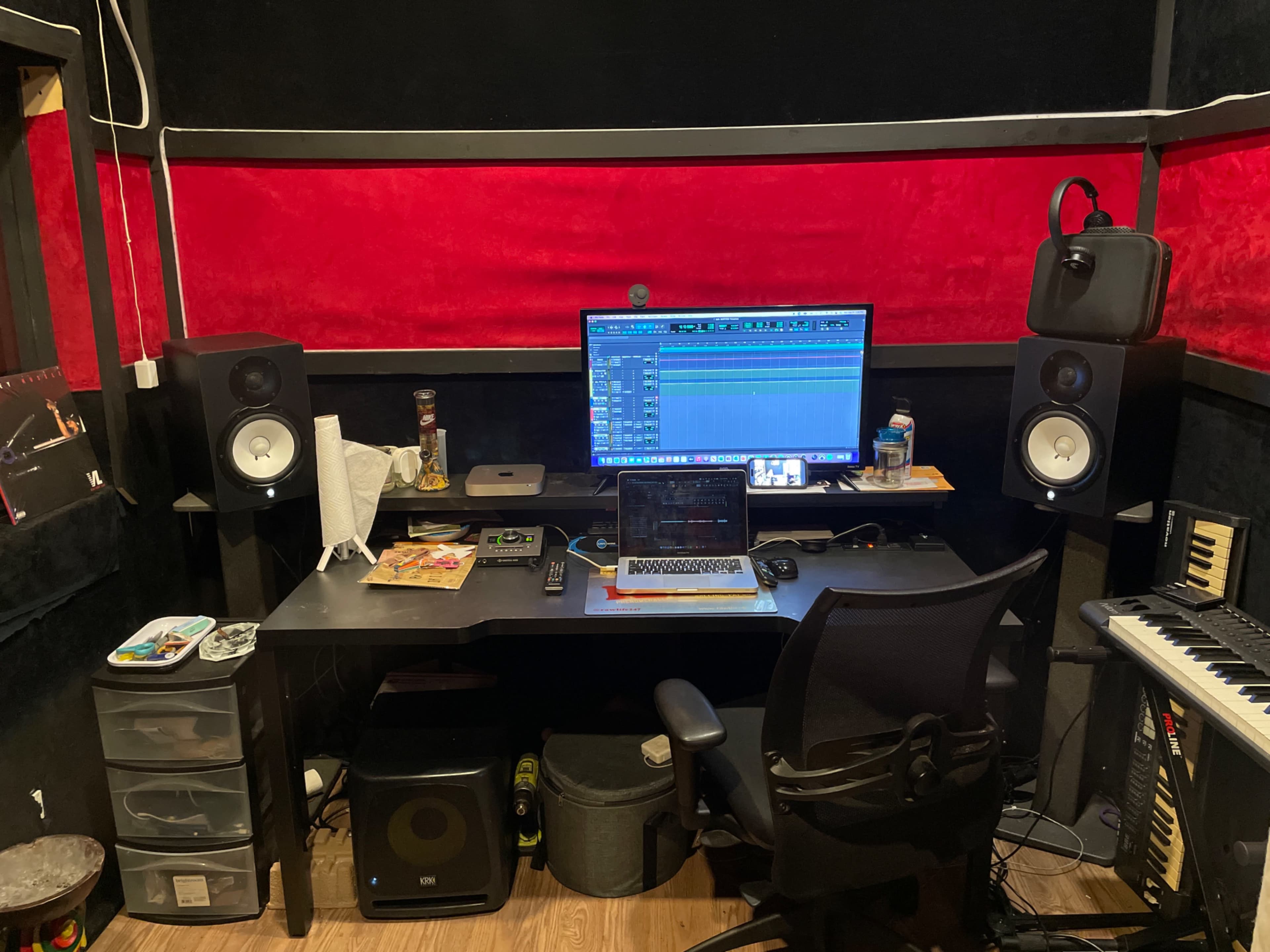 The image shows a music production studio with a computer setup, MIDI keyboard, speakers, and various equipment on a black desk against red and black walls.
