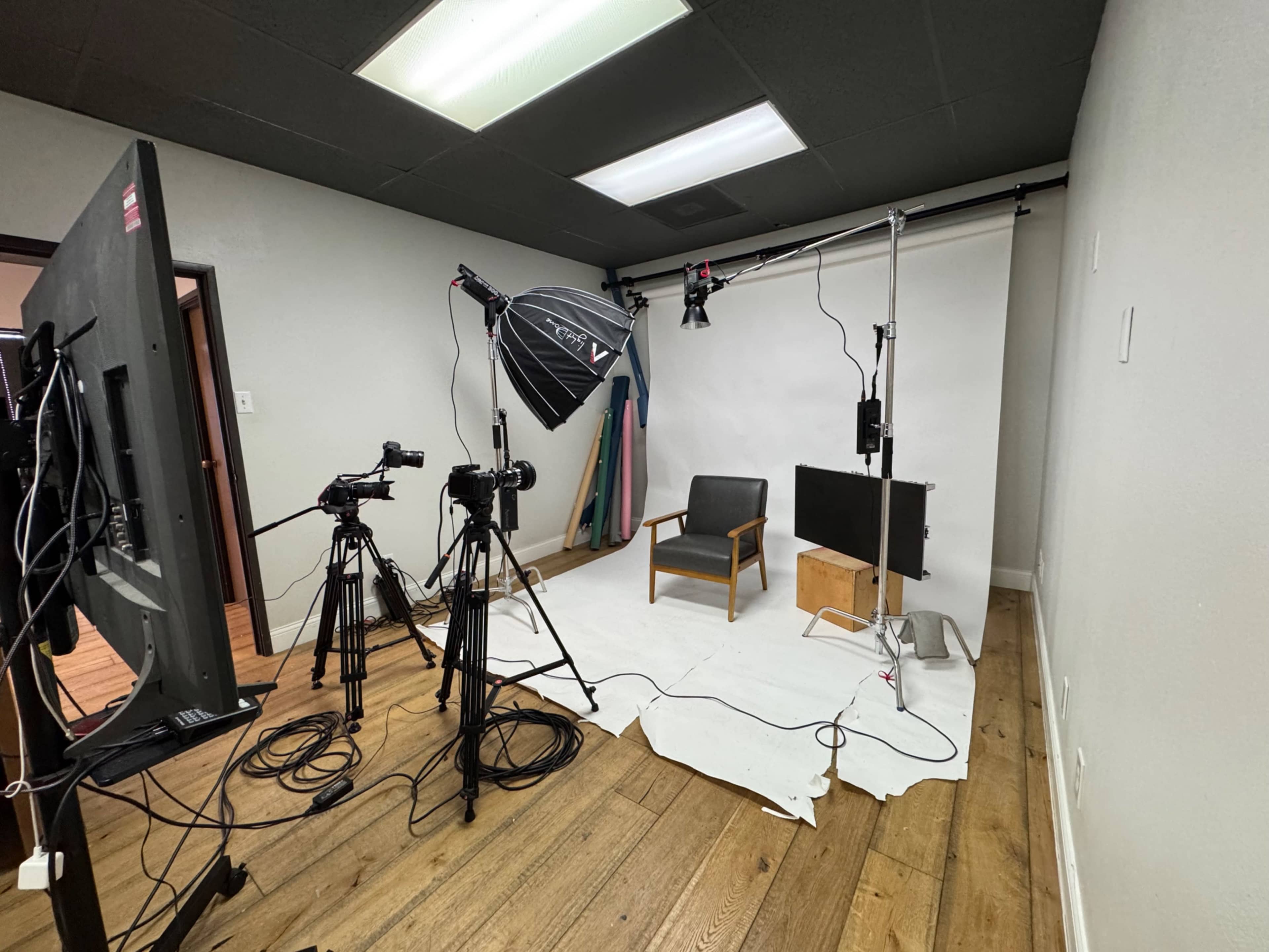 The image shows a small photography studio setup with a chair, lighting equipment, a backdrop, and cameras arranged on tripods.