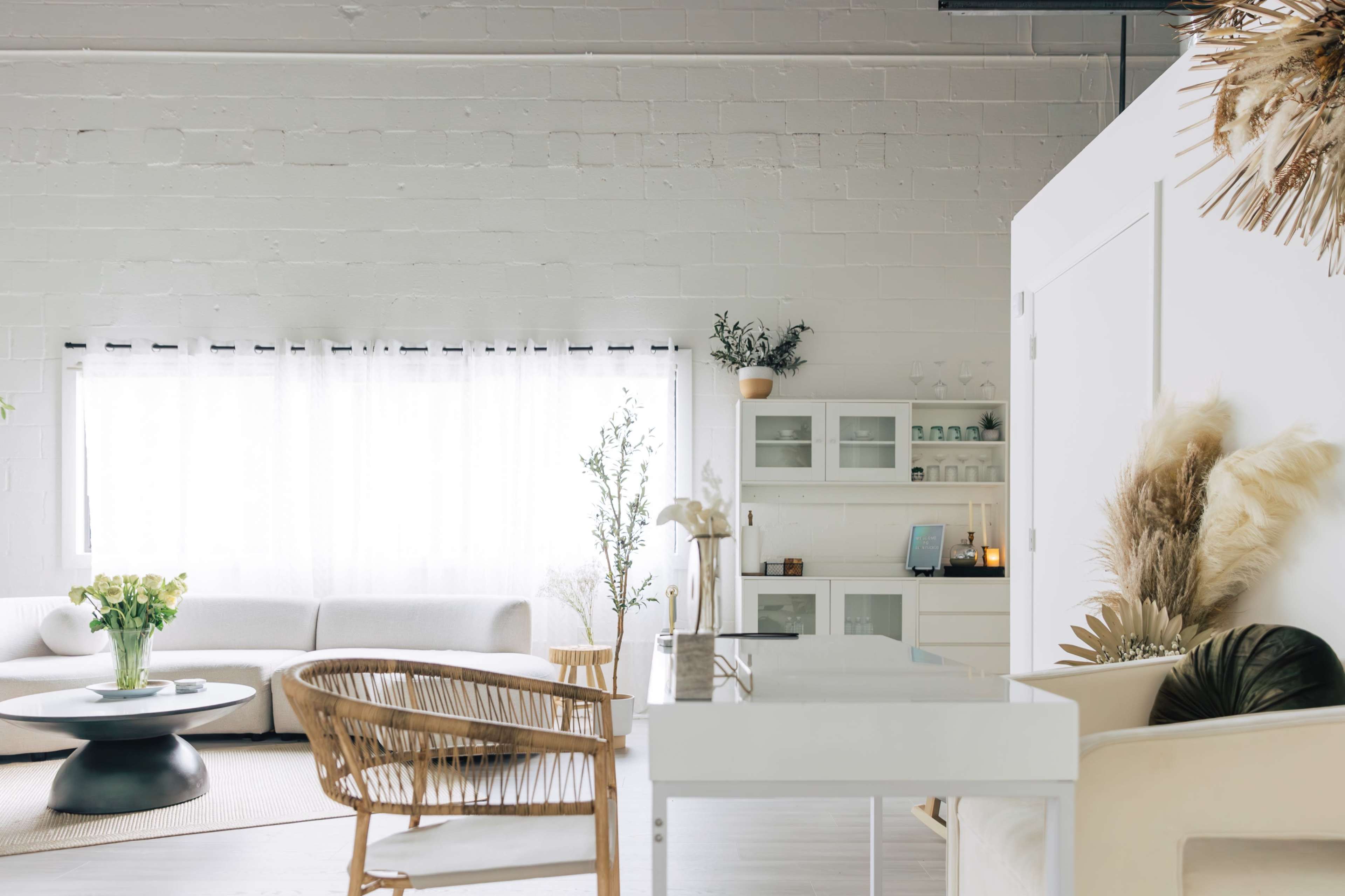 The image shows a bright, minimalist interior featuring a white sofa, a wooden chair, and a sleek white table, with large windows allowing natural light to fill the space.