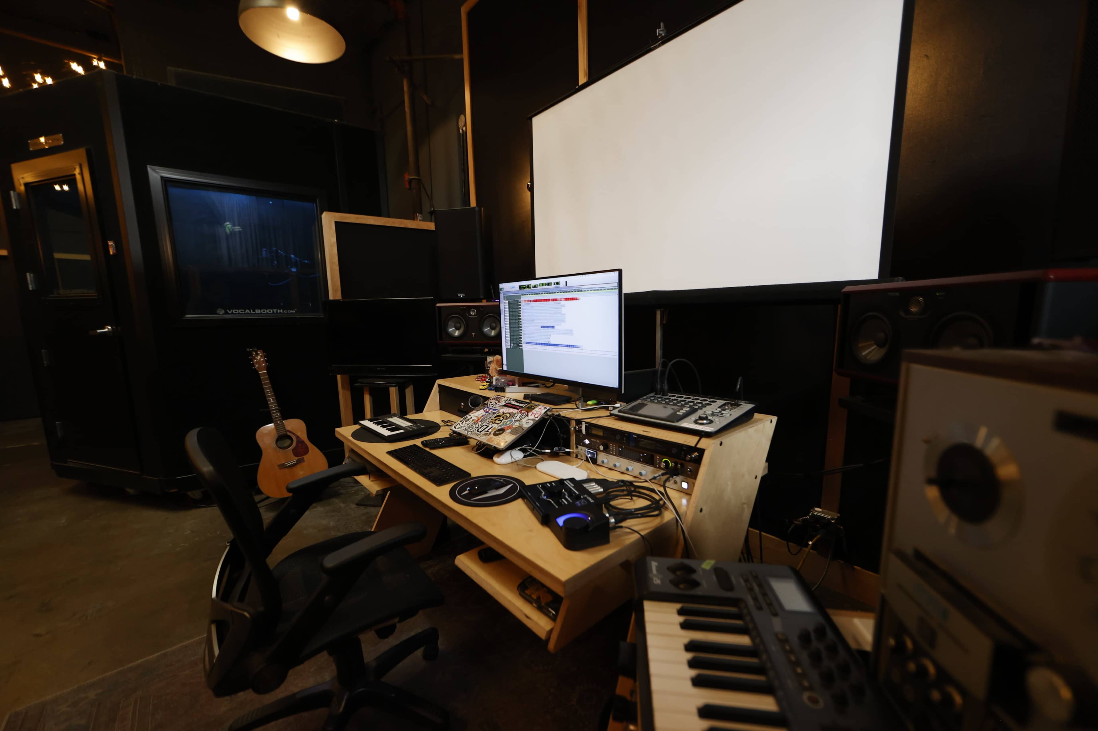 The image shows a music production studio featuring a desk with a computer, audio equipment, an electric guitar, and a large screen.
