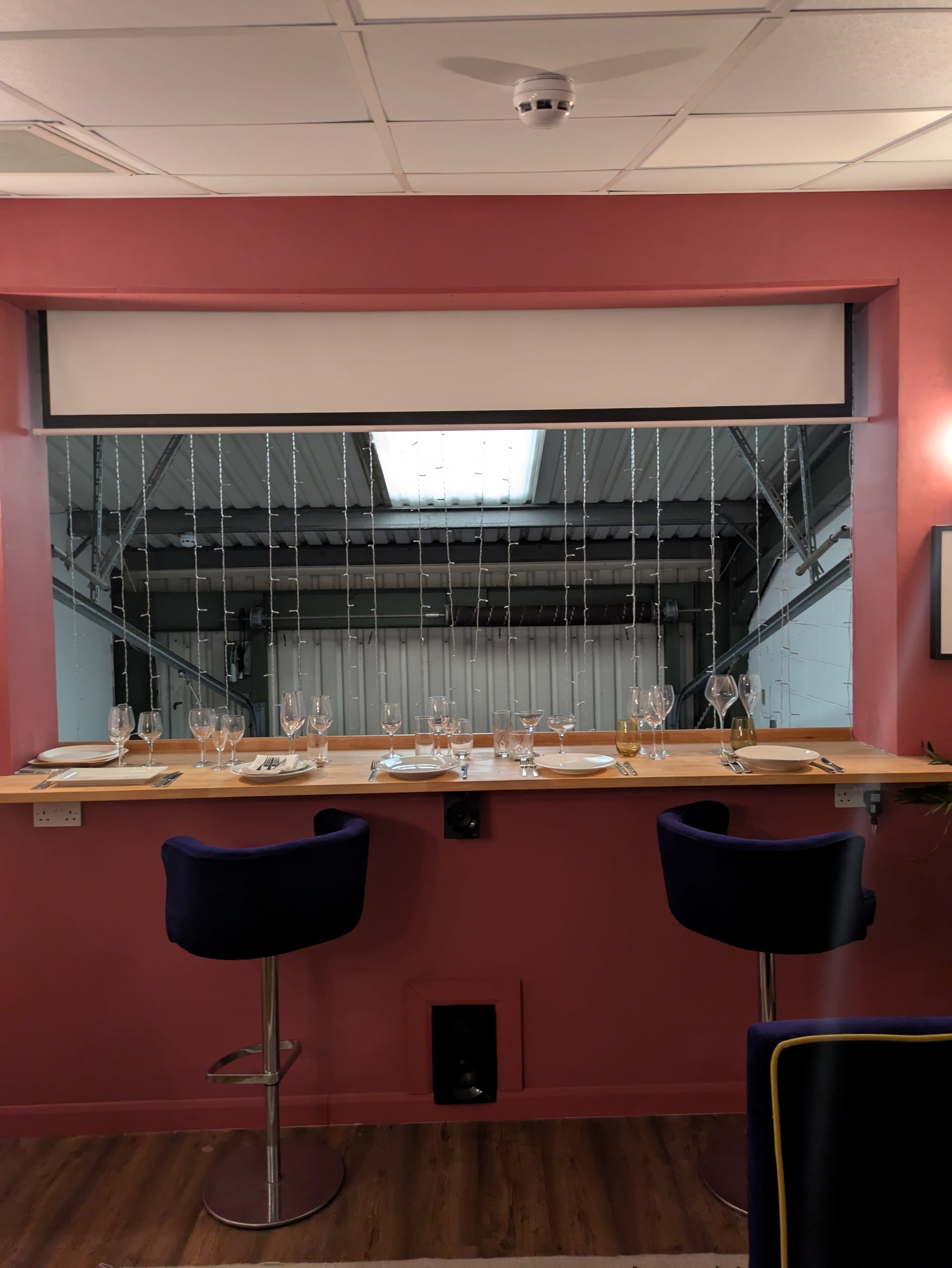 A dining area features a bar with two high stools, set with glassware and cutlery, overlooking an unobstructed view of a skylit industrial space.