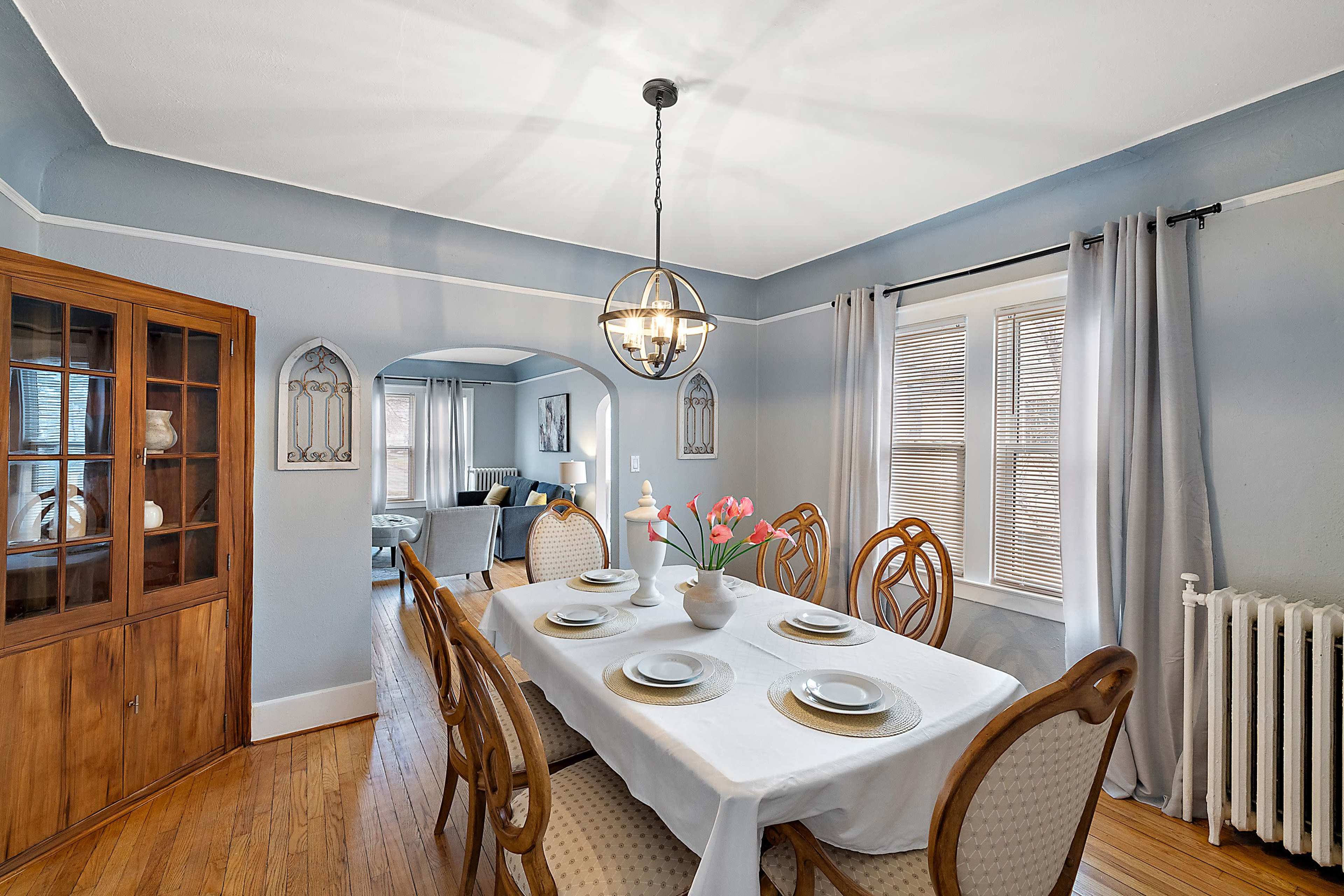 A dining area features a table set for six, surrounded by wooden chairs, with a chandelier overhead and a view into a living room through an arched entry.