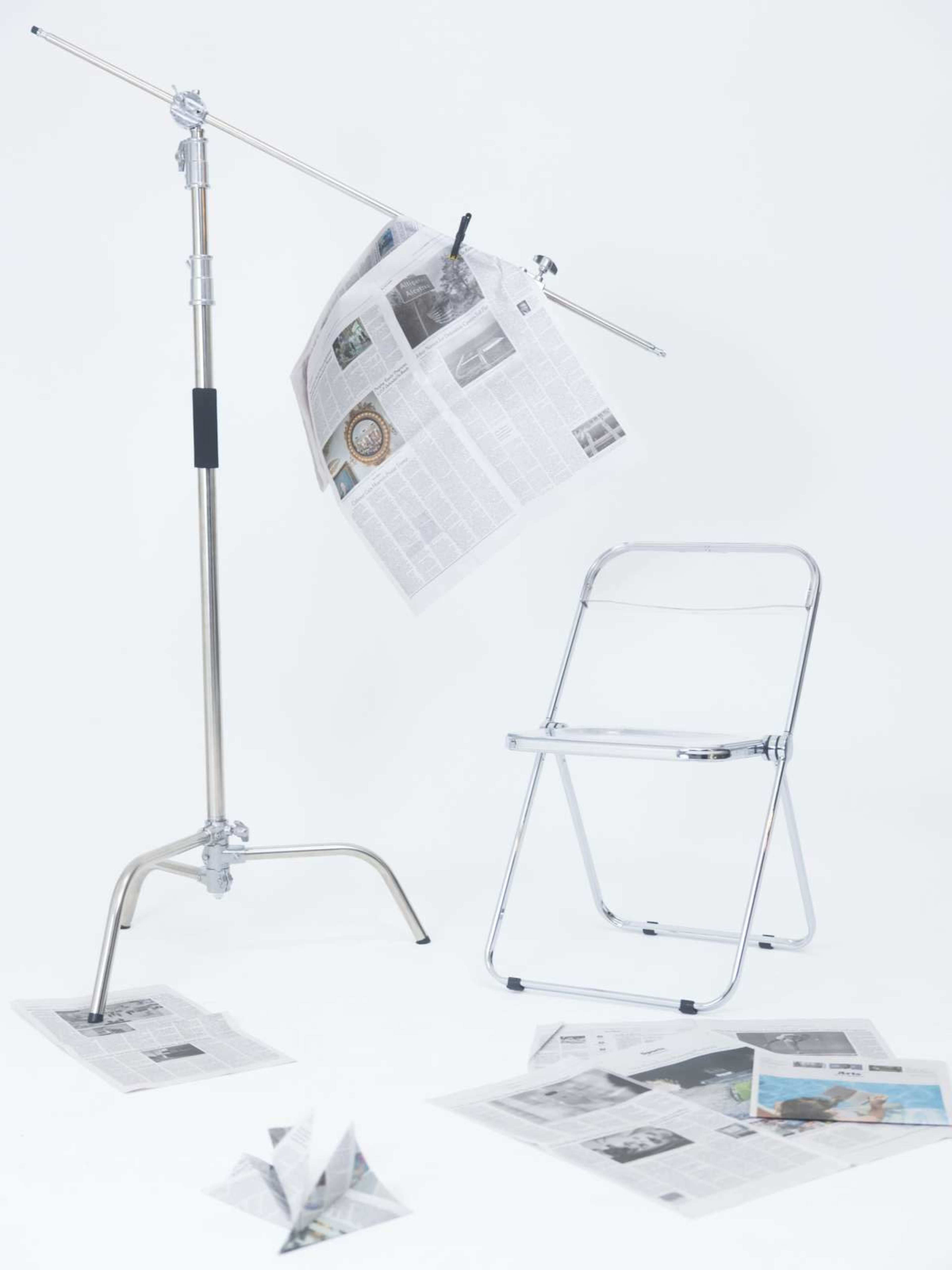 A chrome folding chair stands next to a metal stand holding newspapers, with additional torn newspapers scattered on the floor.