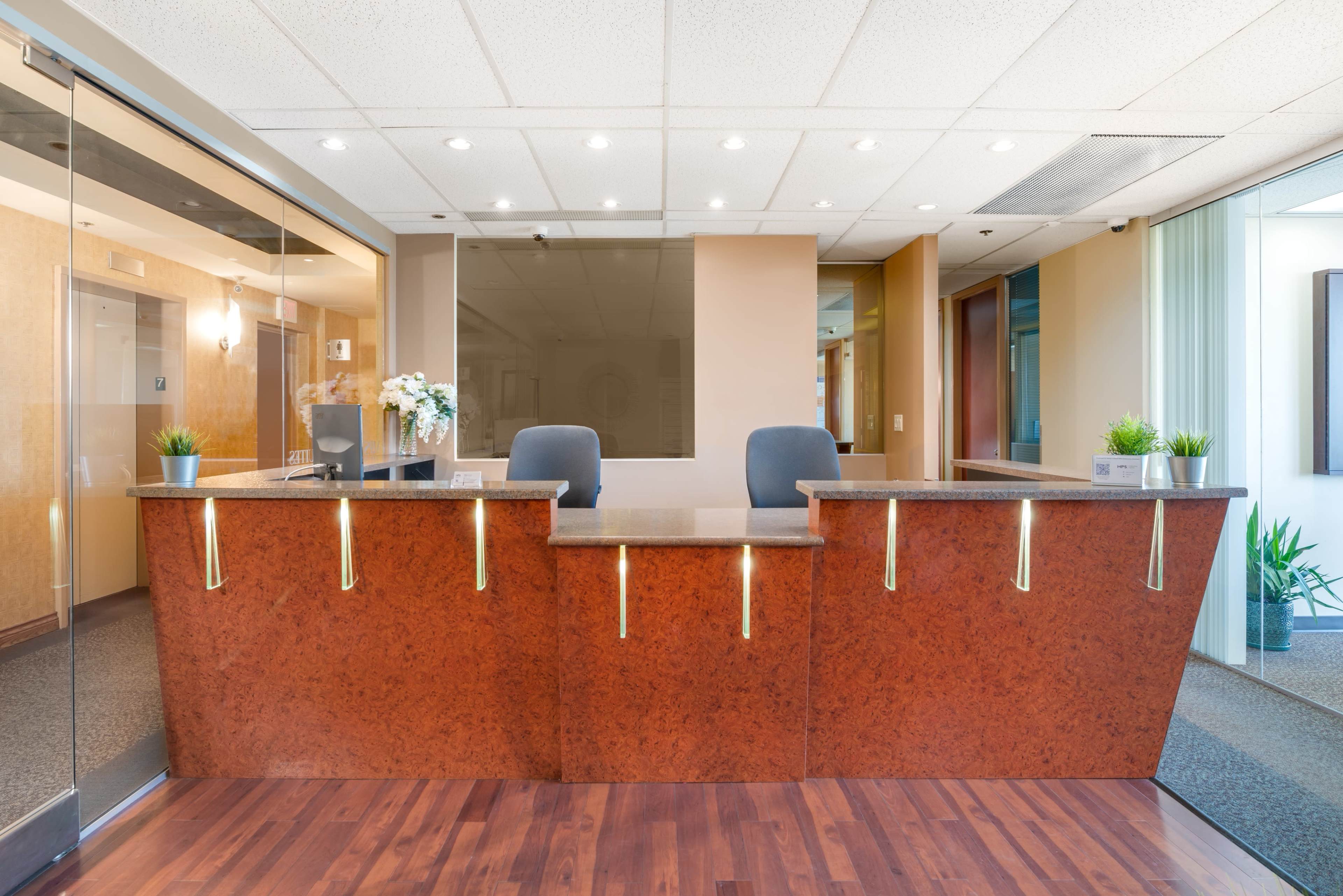 A modern reception desk with a polished surface, two gray chairs, and decorative plants in an office environment.