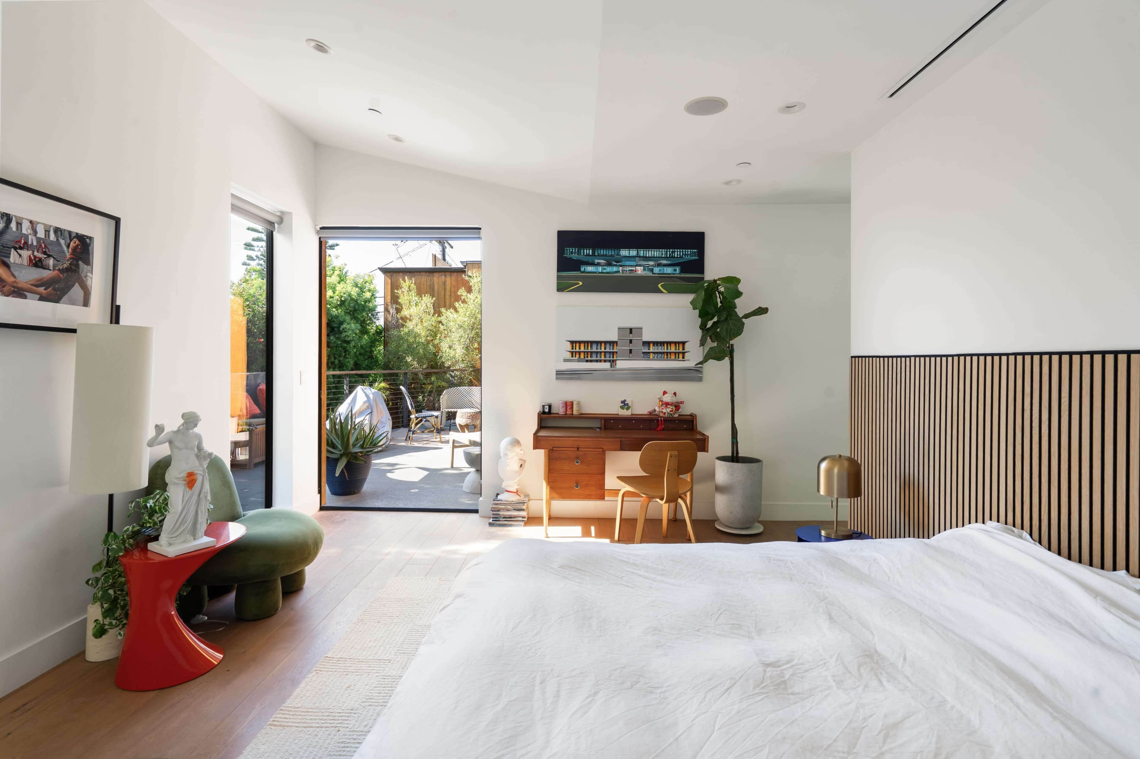 A modern bedroom features a bed, a wooden accent wall, and large windows that open to a small outdoor area with greenery.