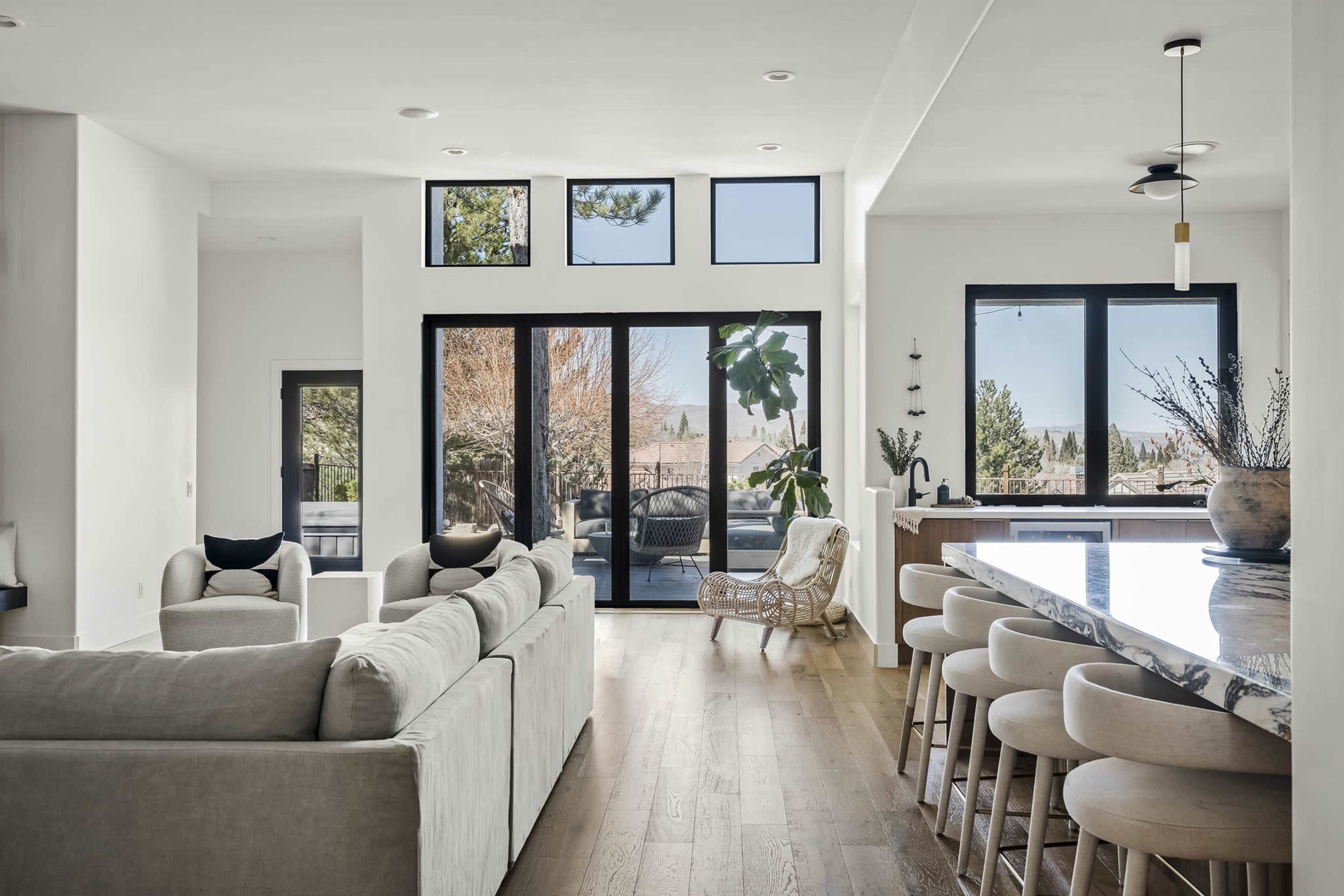 A bright, modern living space featuring a light-colored sofa, a dining area with bar stools, large windows overlooking an outdoor view, and potted plants adding greenery to the decor.