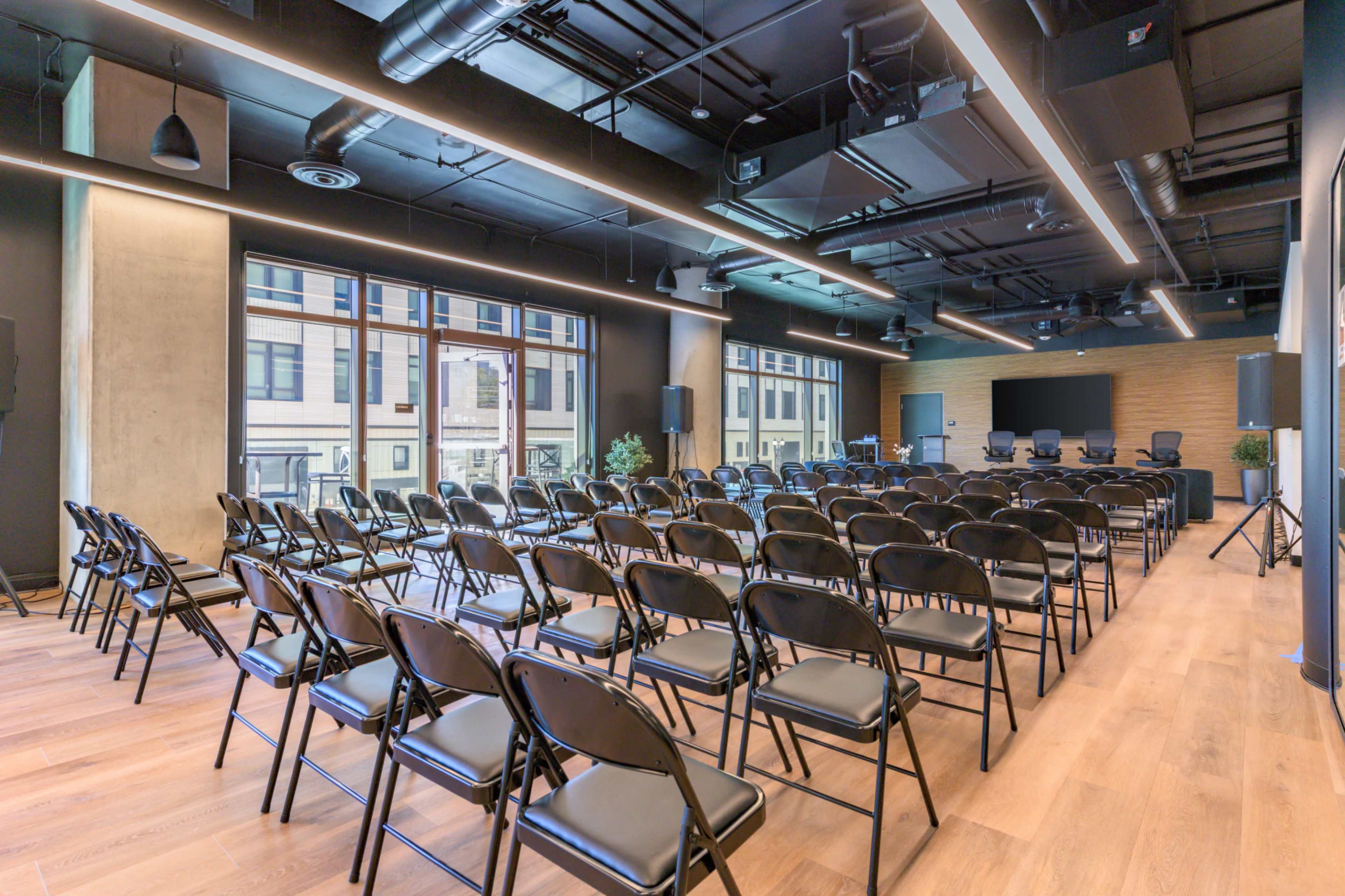 A modern conference room is set up with rows of empty black chairs facing a stage area, featuring large windows that allow natural light.