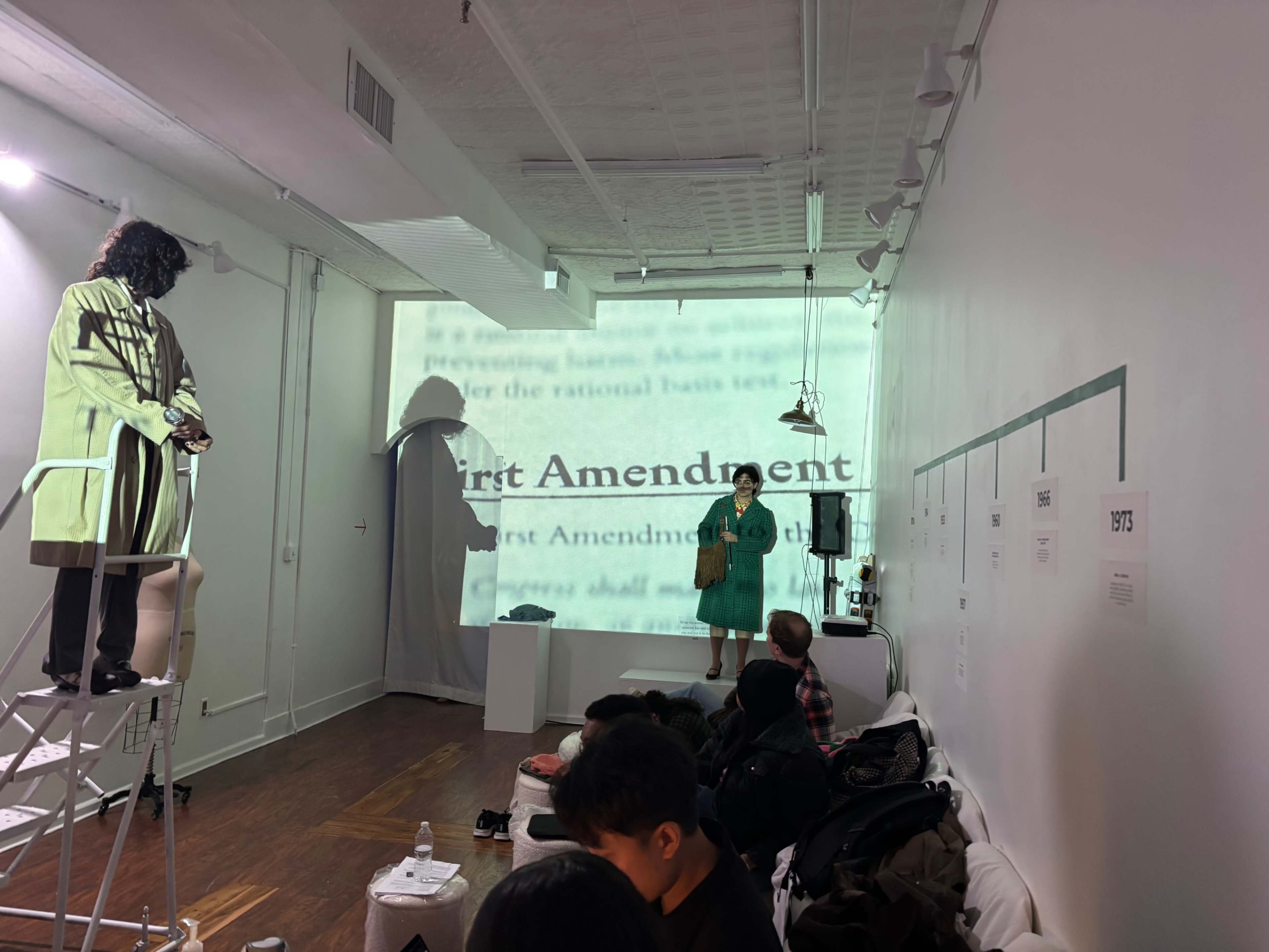 A performer dressed in a green outfit stands on a platform while another figure is projected onto a wall in a gallery setting.