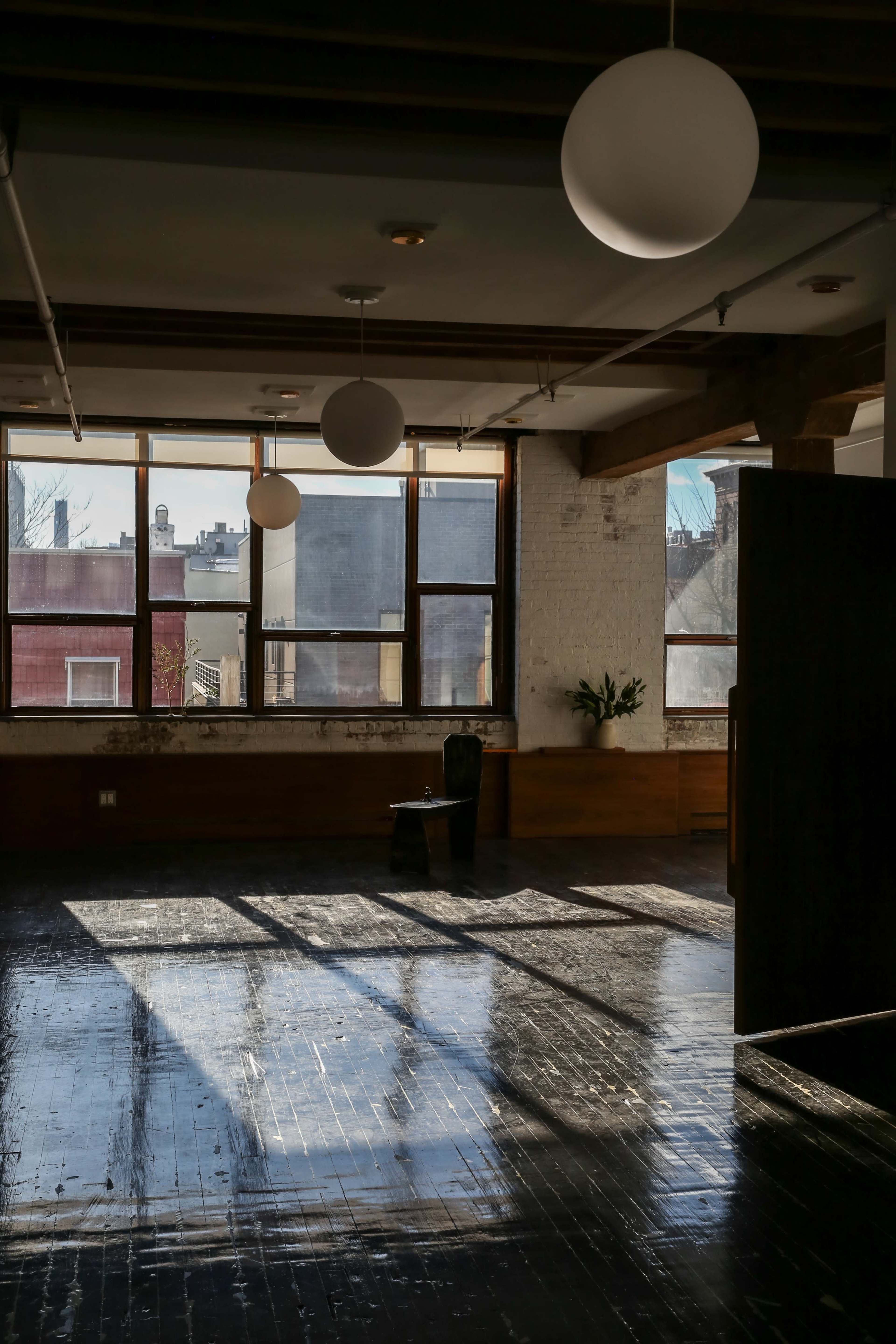 A sunlit room with large windows and a few hanging light fixtures features a solitary chair and shadows on the polished wooden floor.