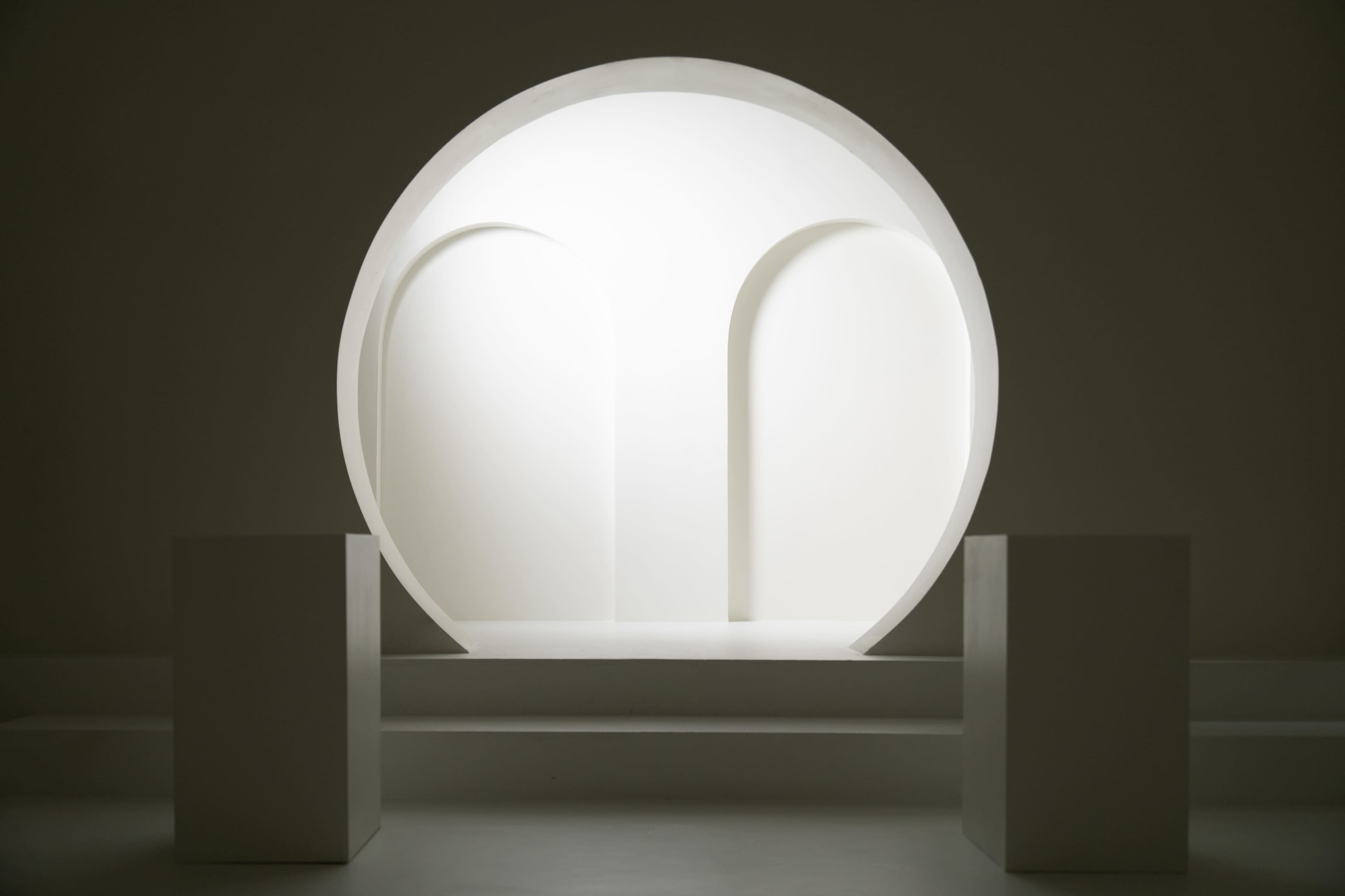 A circular, white architectural structure with arched openings is centered against a minimalist, neutral background.