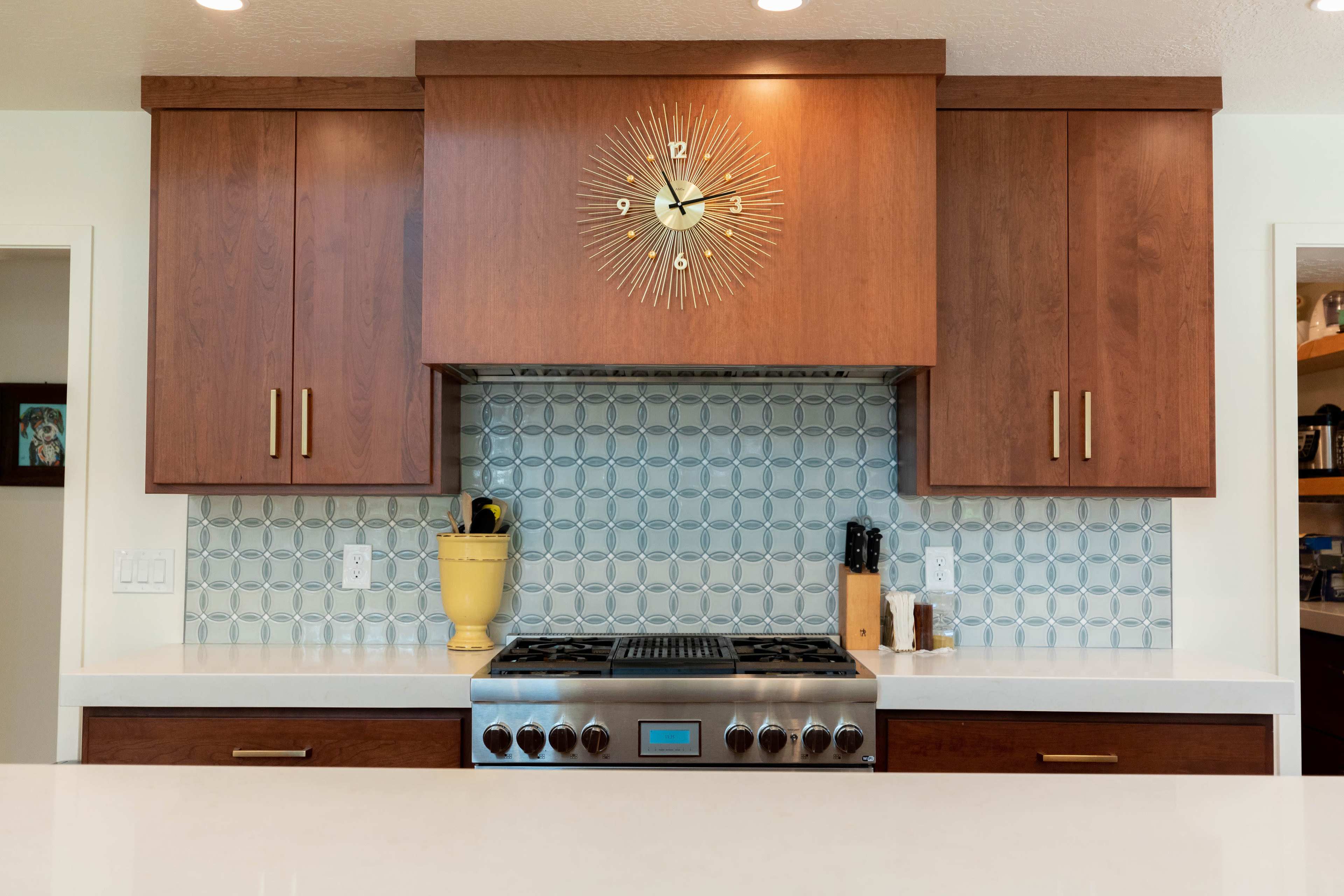A modern kitchen with a gas stove, wood cabinetry, a patterned backsplash, and a decorative wall clock above the range.