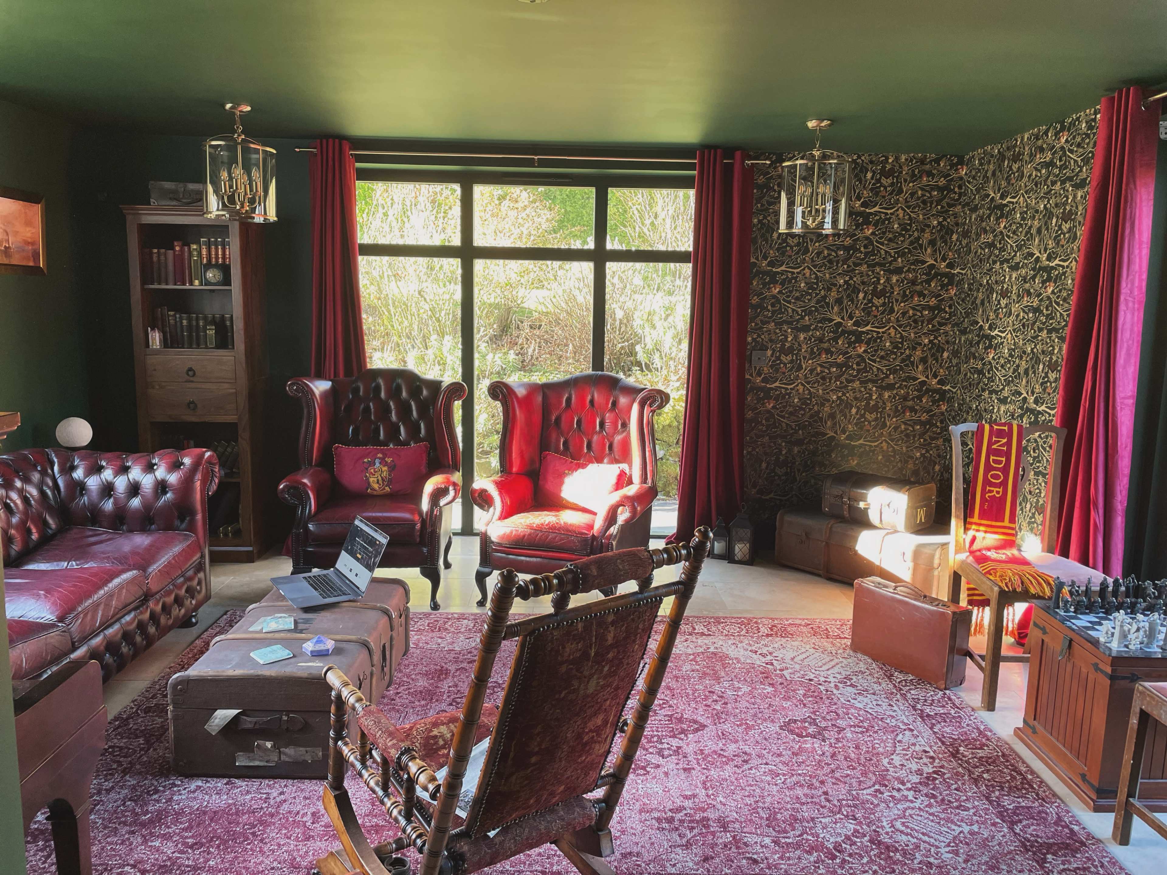 The room features a seating area with two red leather armchairs, a brown sofa, a wooden rocking chair, and a vintage rug, illuminated by natural light from large windows.