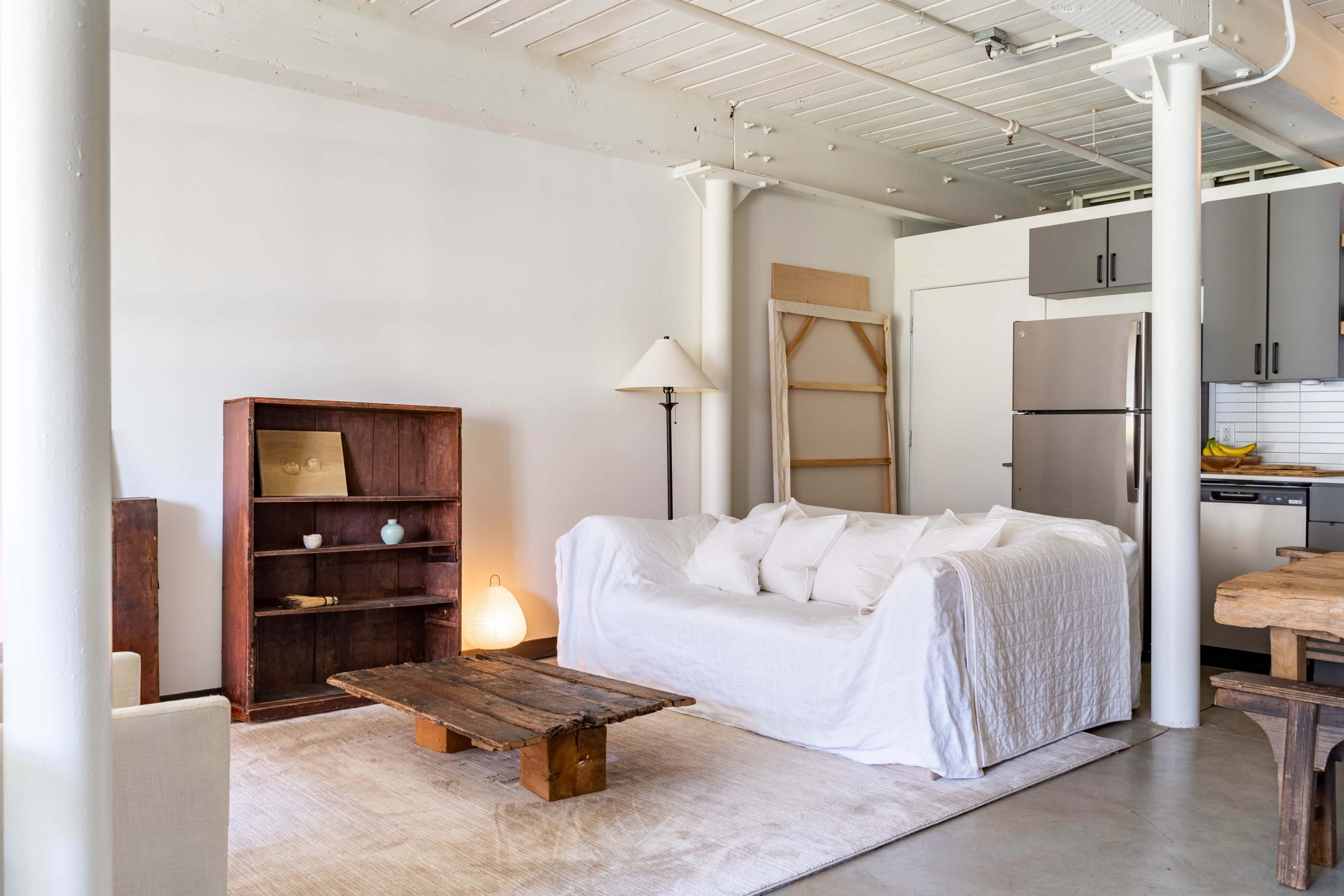 A living room features a white sofa surrounded by wooden furnishings, including a rustic coffee table and a bookshelf, with a lamp and a refrigerator in the background.