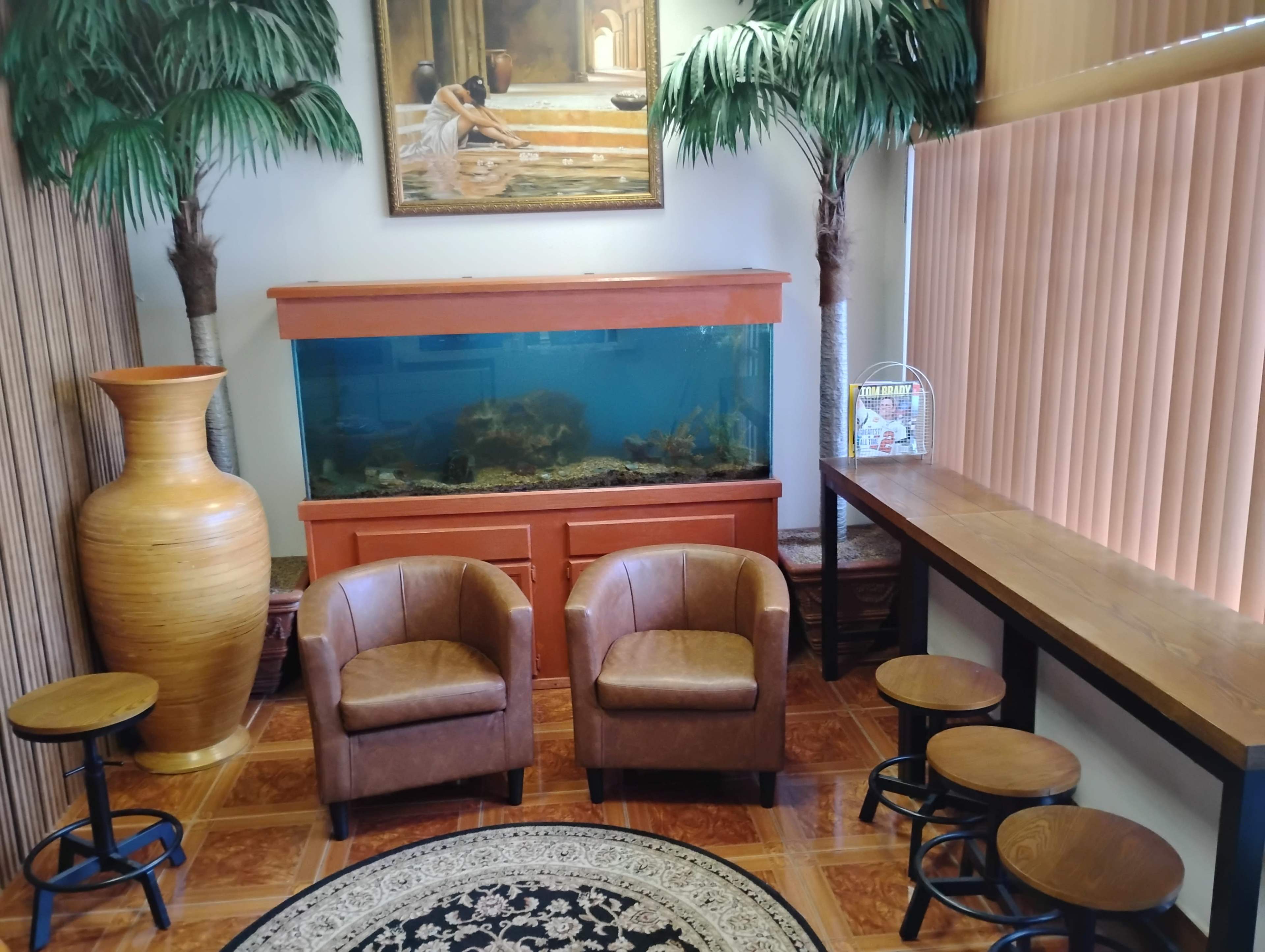 The image shows a cozy waiting area featuring two brown armchairs, a large fish tank, a long wooden table, and decorative plants.