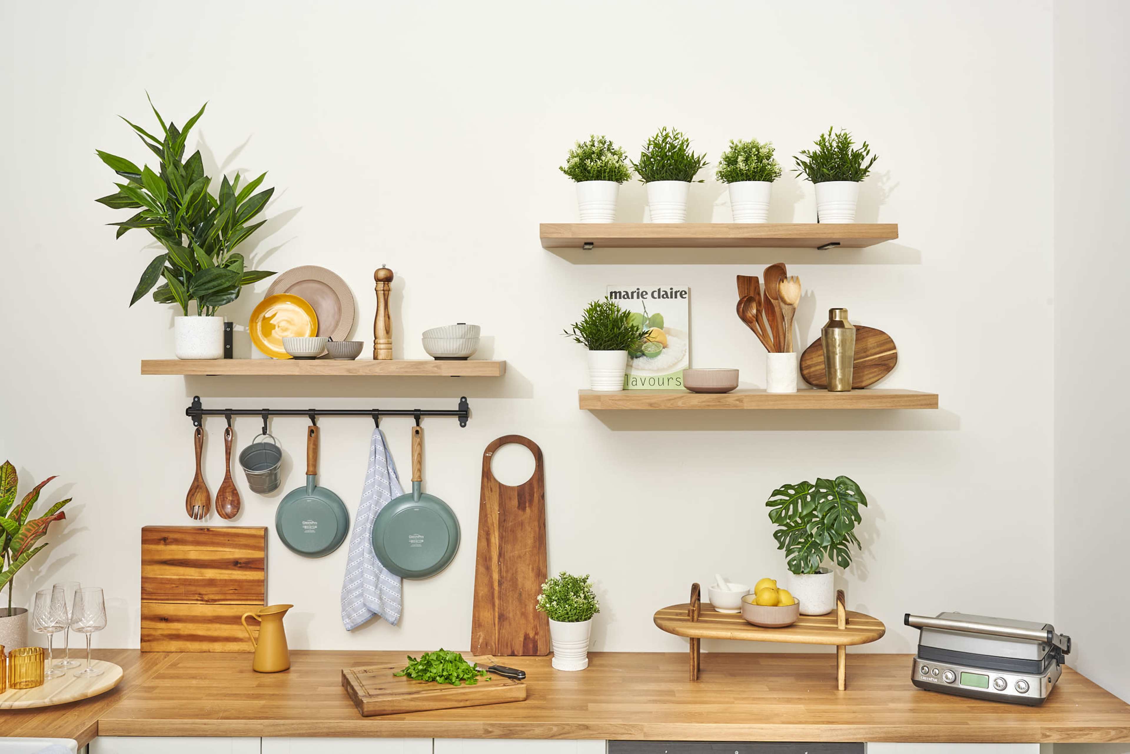 A kitchen scene features open shelves with potted plants, wooden utensils, a cutting board, and cookware arranged neatly on a countertop.